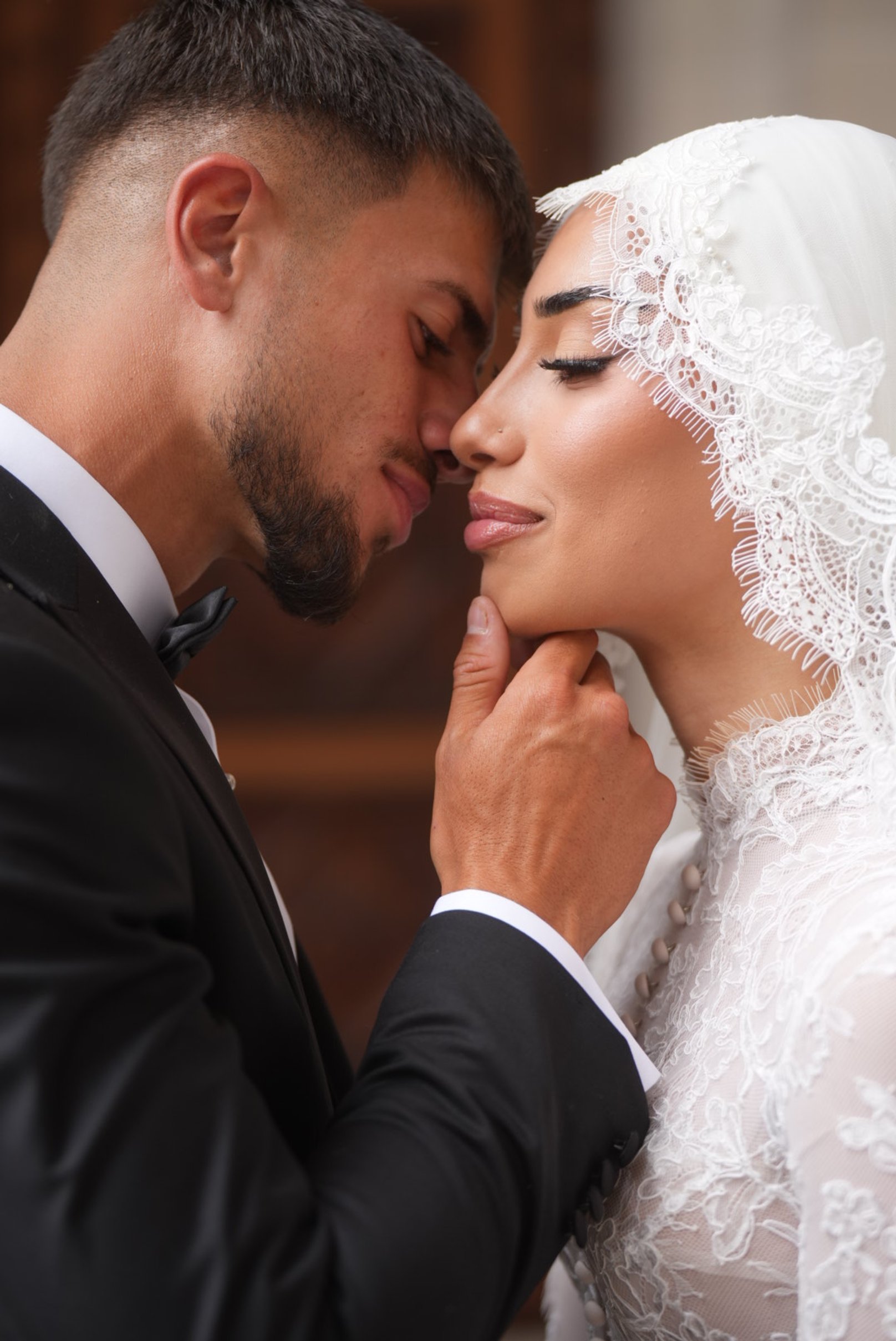 A groom in a black tuxedo gently touching the chin of a bride in a white lace dress, with both showing closed eyes and foreheads touching for an intimate moment.