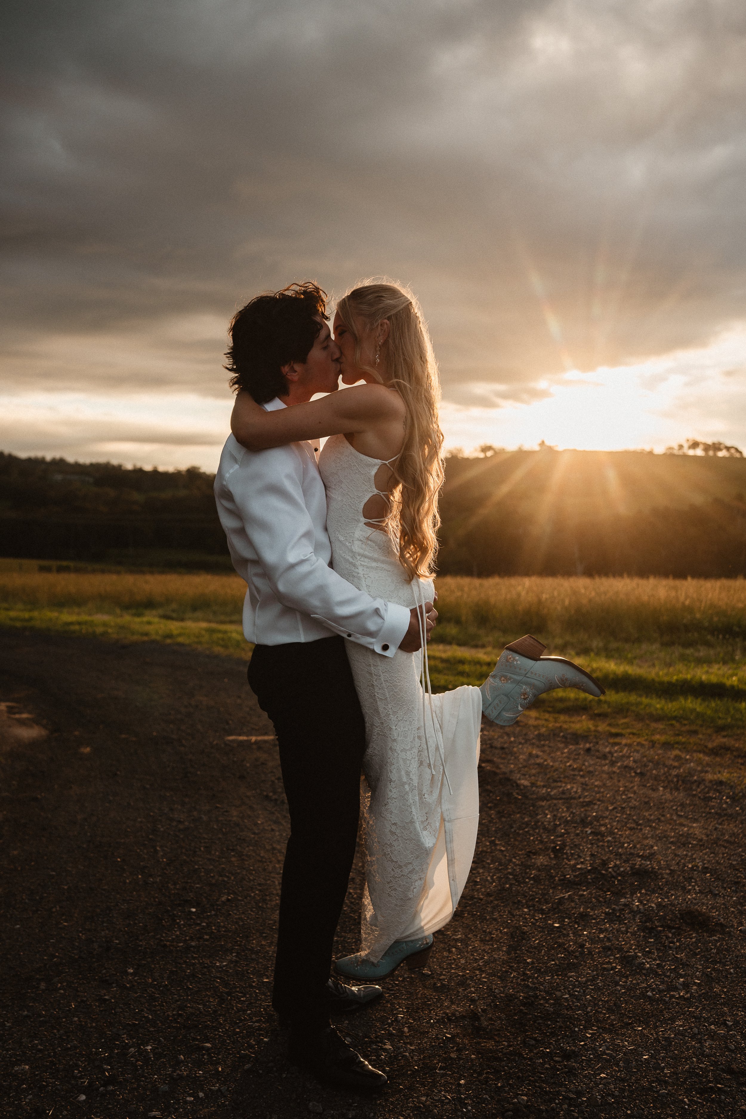 A couple dressed in wedding attire, kissing outdoors at sunset, with the groom lifting the bride's leg.