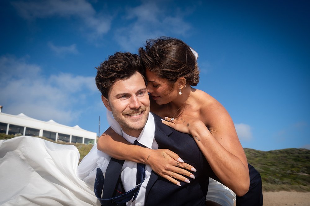 A woman in a wedding dress hugging a man or husband in a suit outdoors against a blue sky, possibly beach