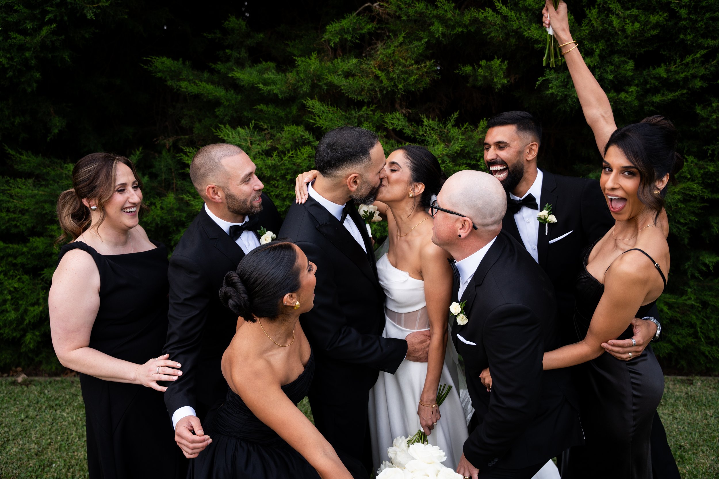 Group of people in formal attire celebrating wedding, with a couple kissing in the center surrounded by friends.