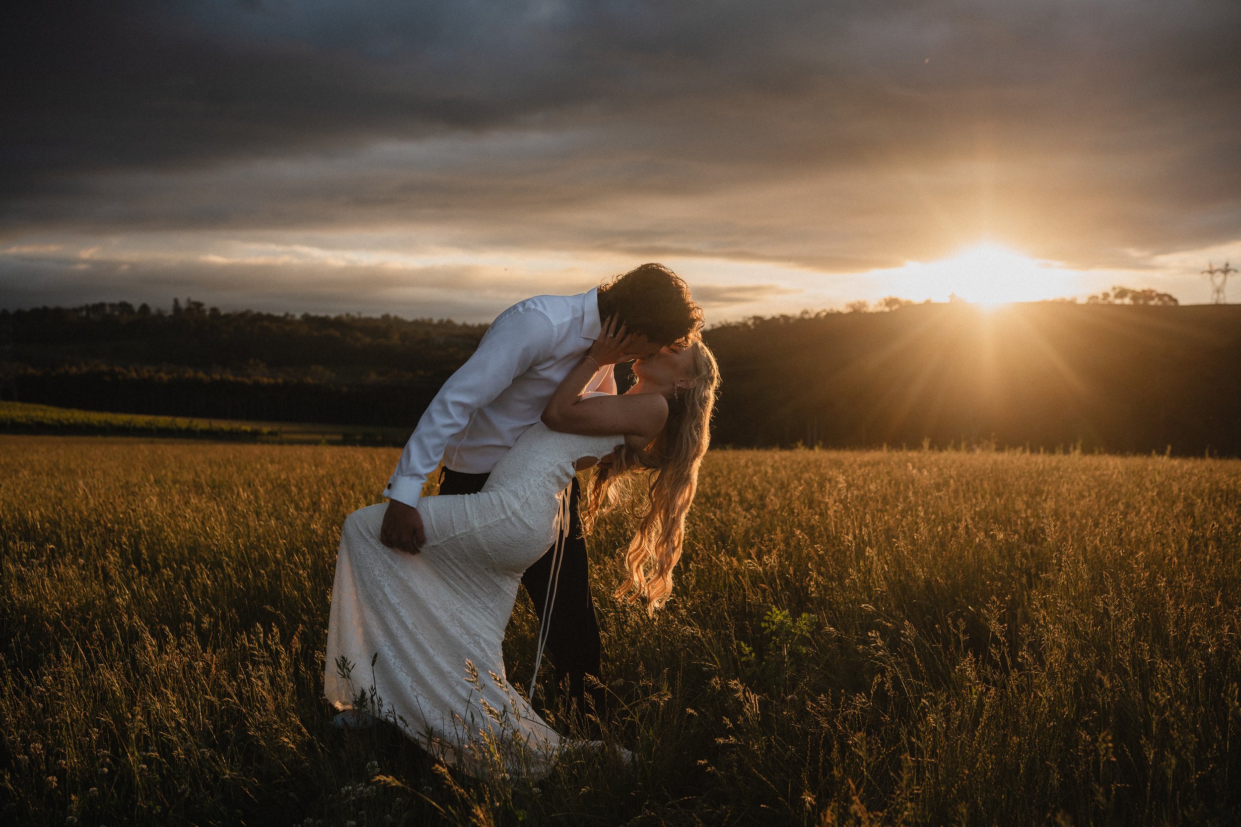 A couple in wedding attire sharing a kiss in a field at sunset.