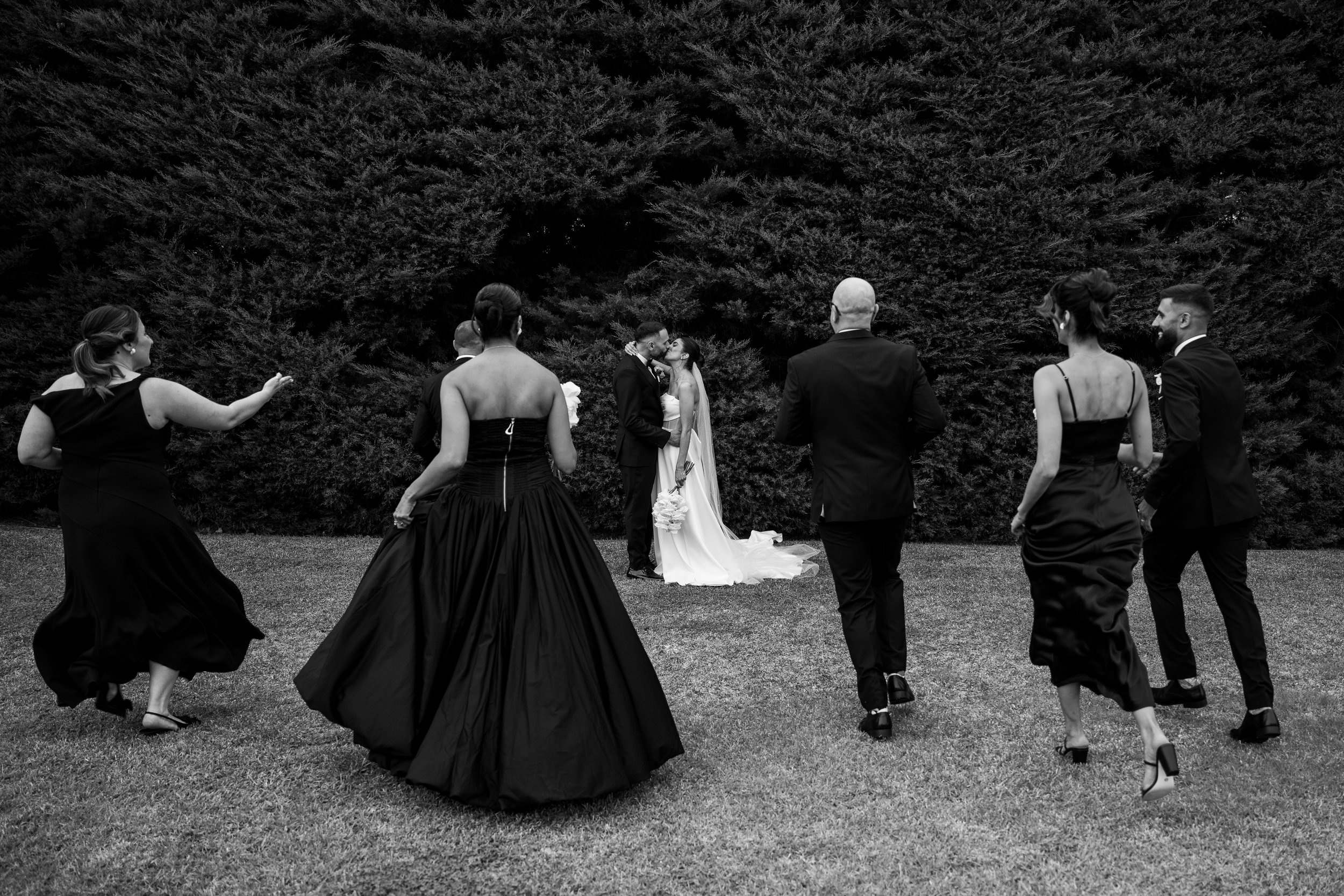 A group of people dressed in formal attire, dancing outside, with a bride and groom in the center, surrounded by a large hedge.