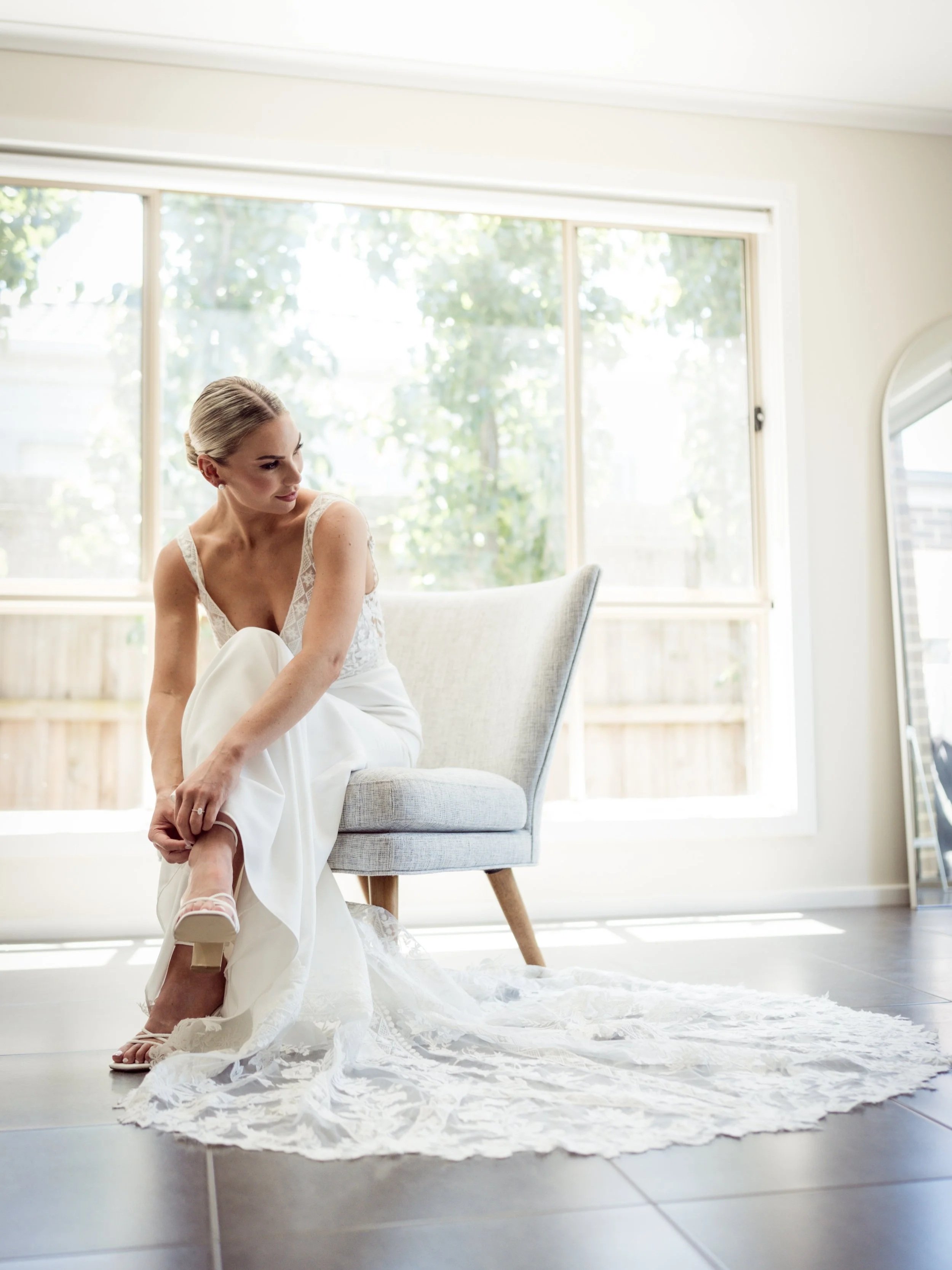 A bride in a white wedding dress sitting on a light gray armchair, getting ready on the morning of her wedding, casually adjusting her shoe, in a room with large windows and sunlight filtering through trees outside.