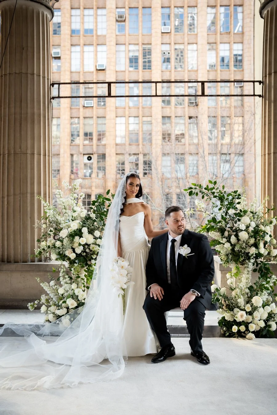 Bride in a white wedding dress and veil standing beside groom in a black suit, seated on a bench with floral arrangements, in front of a large window with a cityscape background.