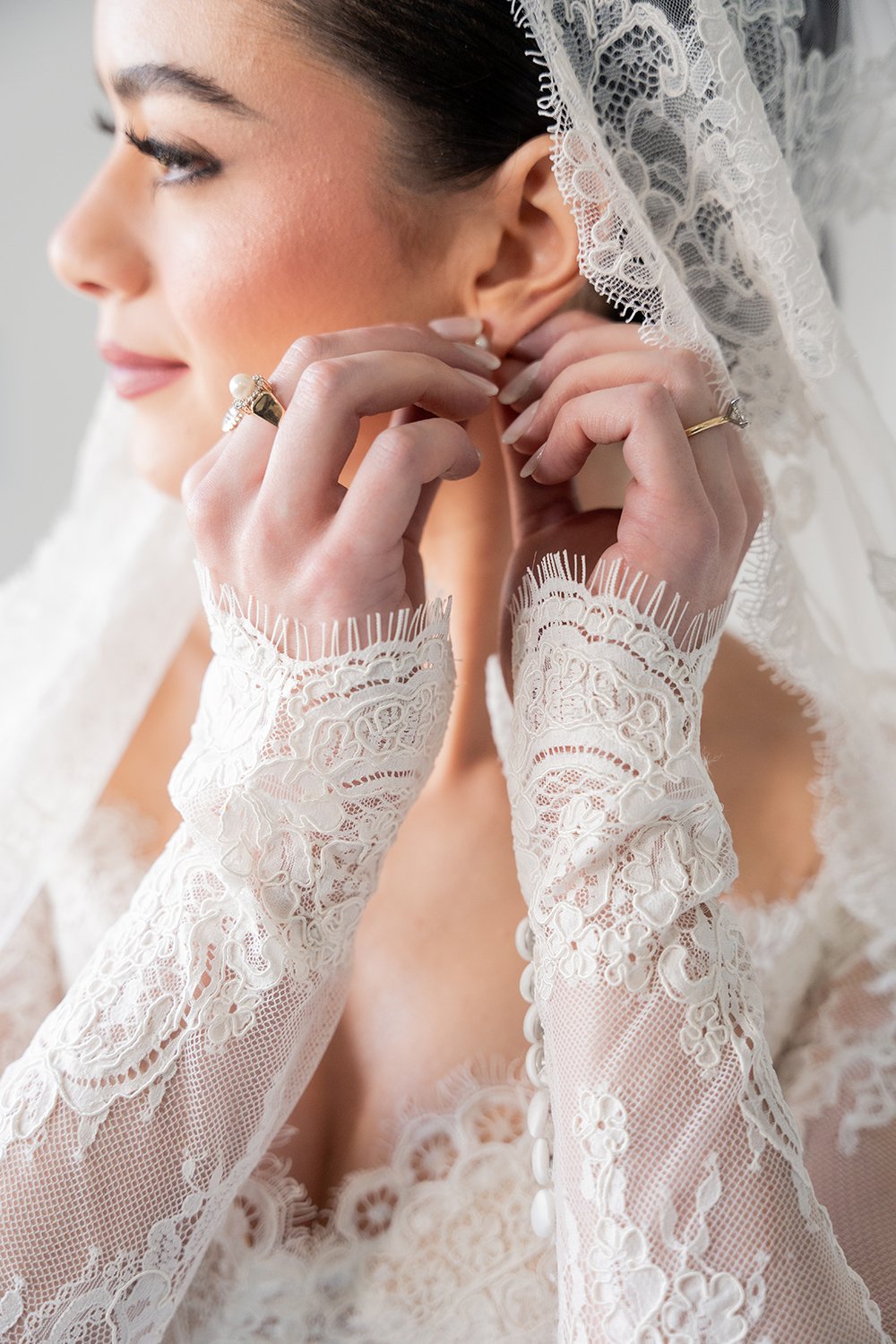 Close-up of a bride putting on earrings, wearing lace gloves and a lace veil.