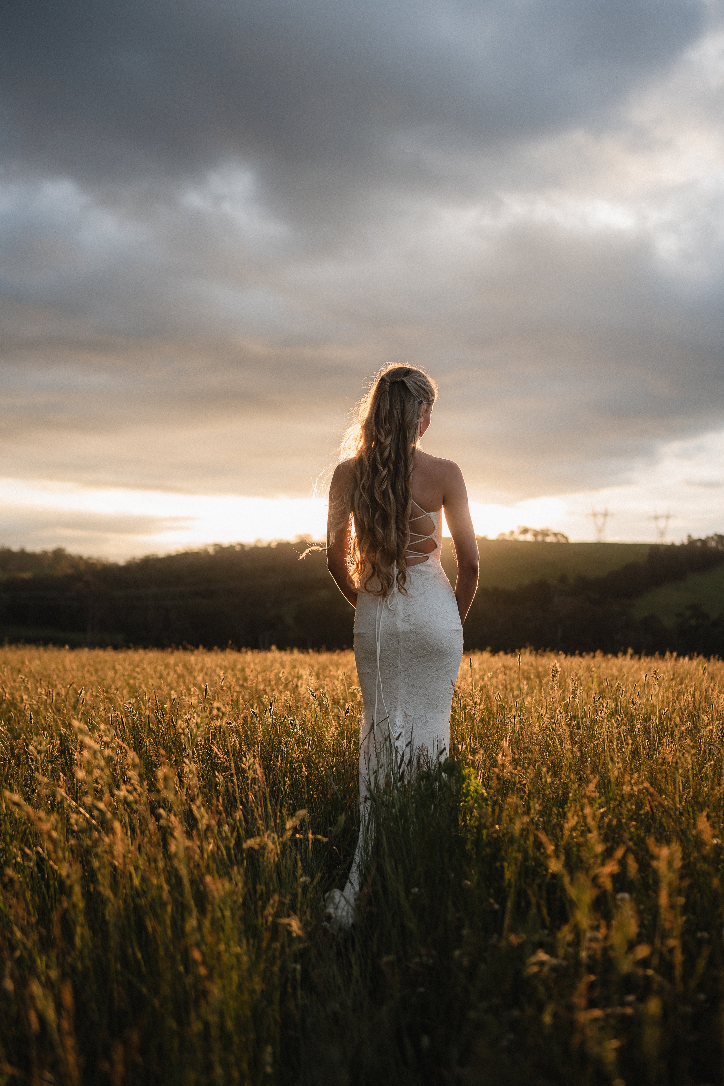 A woman in a white lace wedding dress with open lace-up back stands in a grassy field at sunset, facing away from the camera, with clouds in the sky.