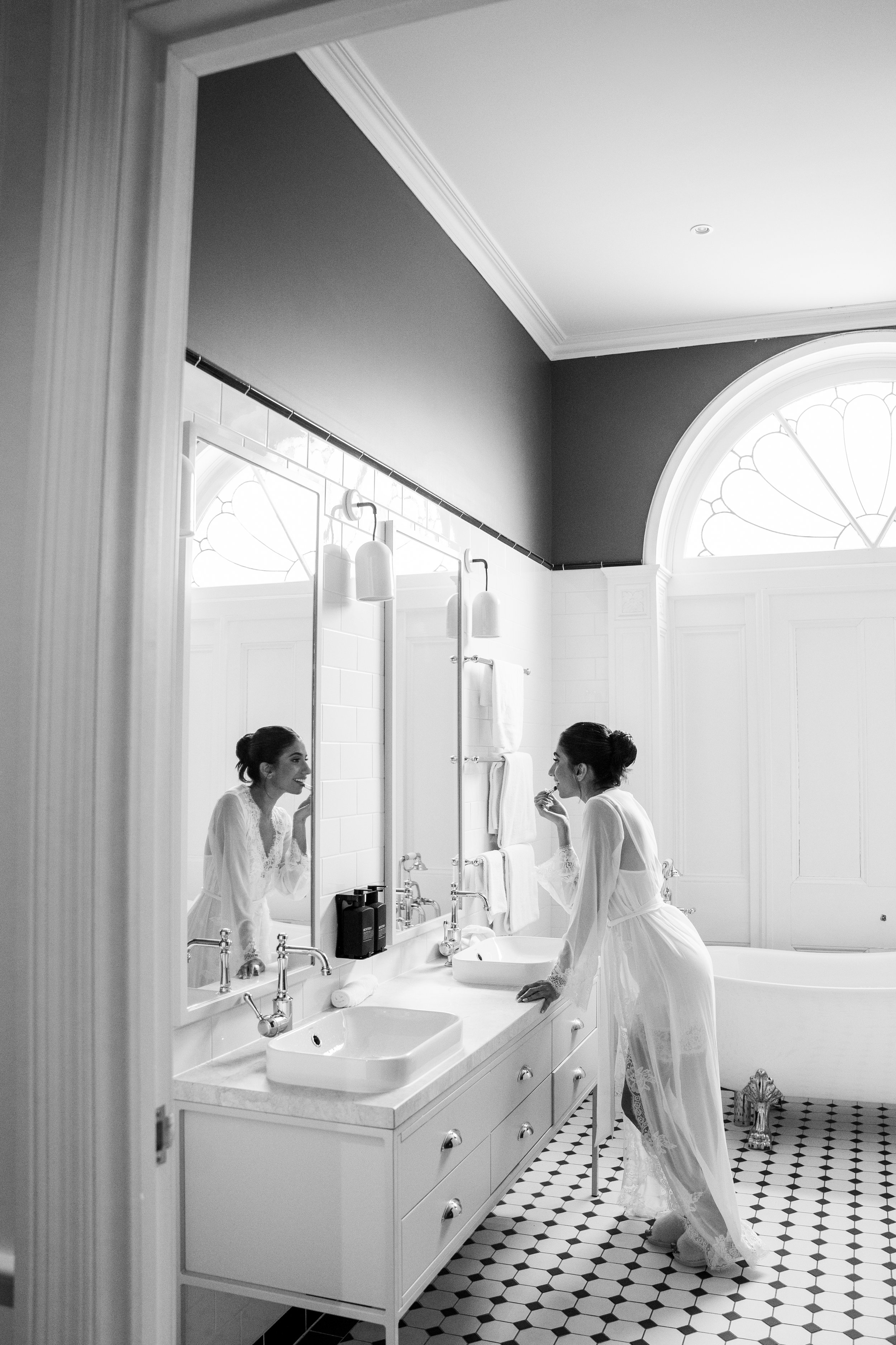 A woman in a sheer white dressing gown is standing at a bathroom vanity, applying lipstick while looking in a mirror, with a clawfoot bathtub and decorative window in the background.