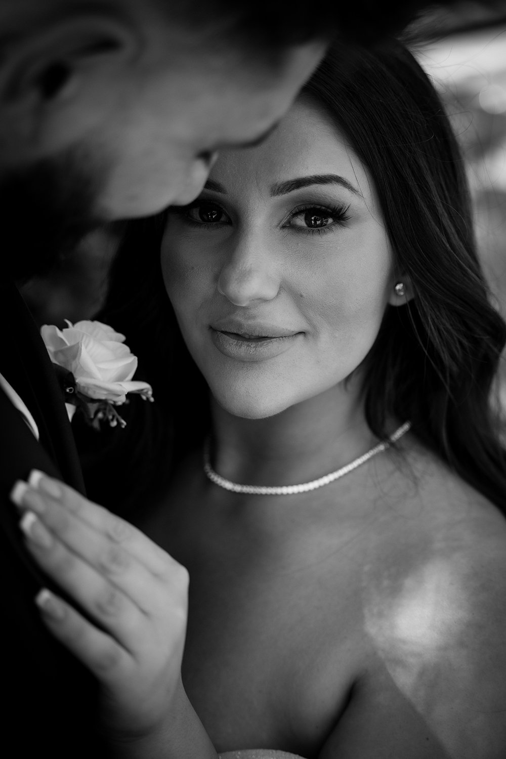A black and white close-up photo of a woman with dark hair looking at the camera, with a man's face slightly visible near her, wearing a suit with a boutonniere.