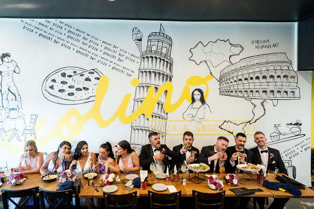 A group of people, dressed formally, sitting at a long table with plates of food and bouquets of flowers, celebrating in a restaurant with a large mural of famous Italian landmarks and illustrations on the wall behind them.
