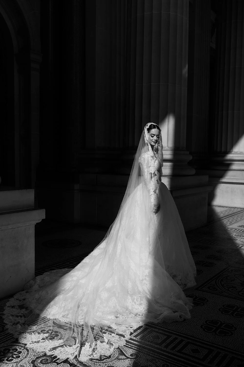 A bride in a wedding dress with a long train standing in a grand, architecturally detailed hall with tall columns. She is smiling and looking down, with a veil over her head, illuminated by a shaft of light.
