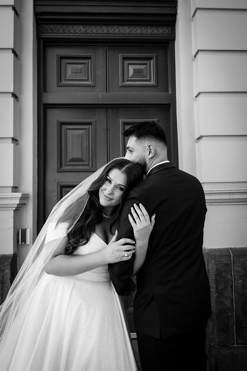 A black-and-white photo of a bride and groom embracing in front of a large wooden door. The bride is smiling and leaning her head on the groom's shoulder, with her hands resting on his arm, while the groom is facing away from the camera.