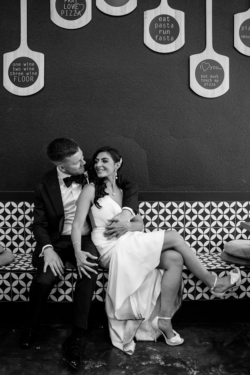 A black-and-white photo of a couple in formal attire sitting on a patterned bench inside a restaurant or cafe. The man, wearing a tuxedo, is smiling and looking at the woman. The woman, wearing a white dress and high heels, is smiling back and leanin