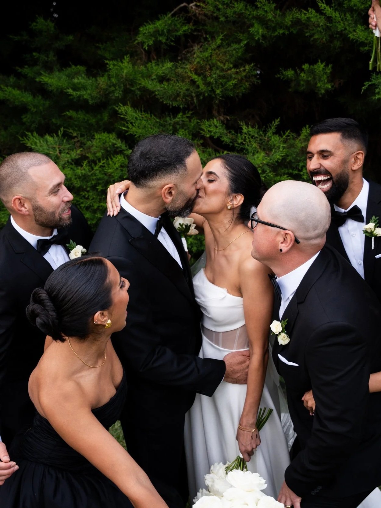 Couple kiss while surrounded by friends in formal attire at a wedding celebration outdoors.
