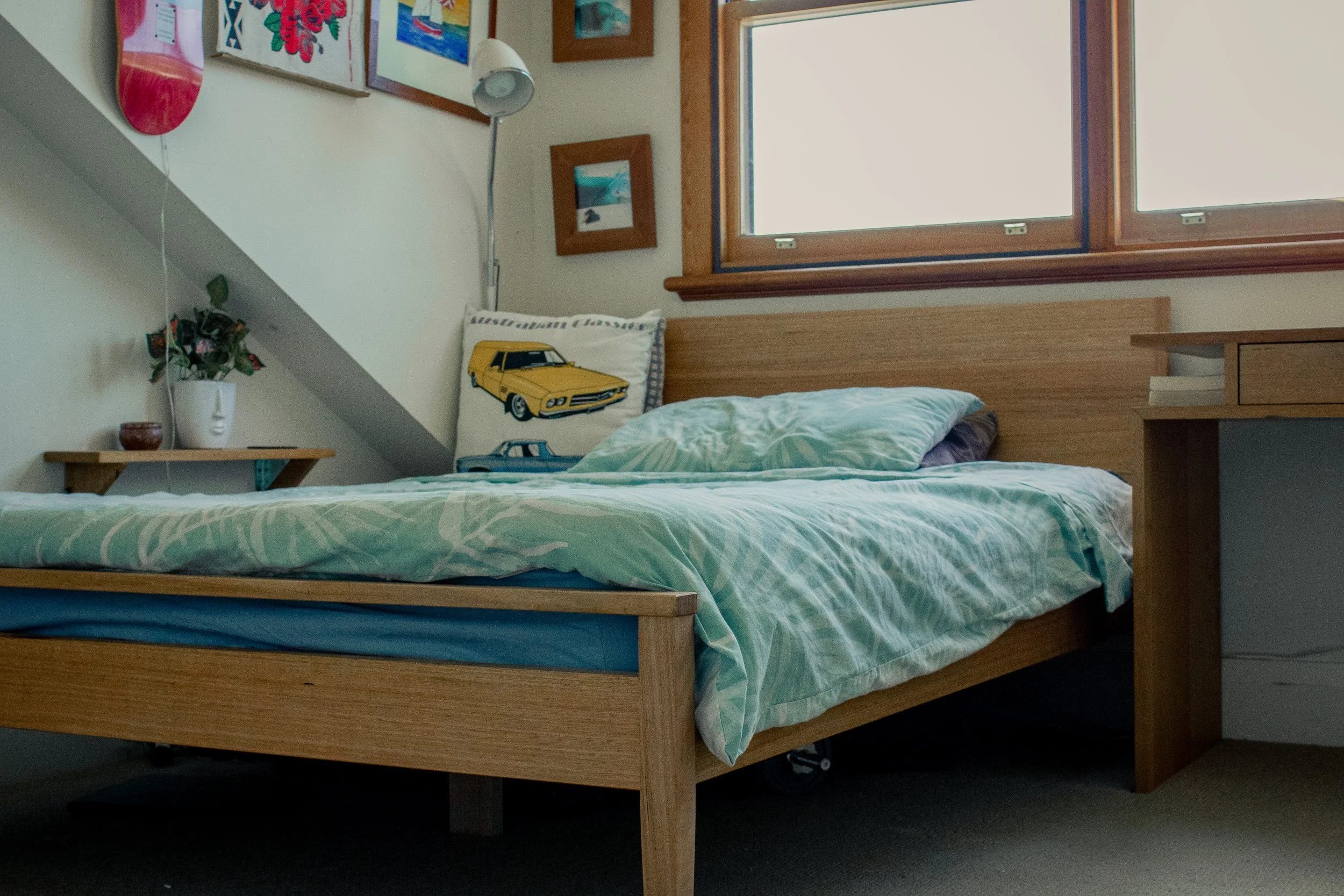A neatly made bed with a wooden frame, a green patterned duvet, and a pillow featuring a yellow cartoon car design. To the left, a small wooden nightstand holds a white vase with artificial flowers and a small brown container. The wall above the bed 