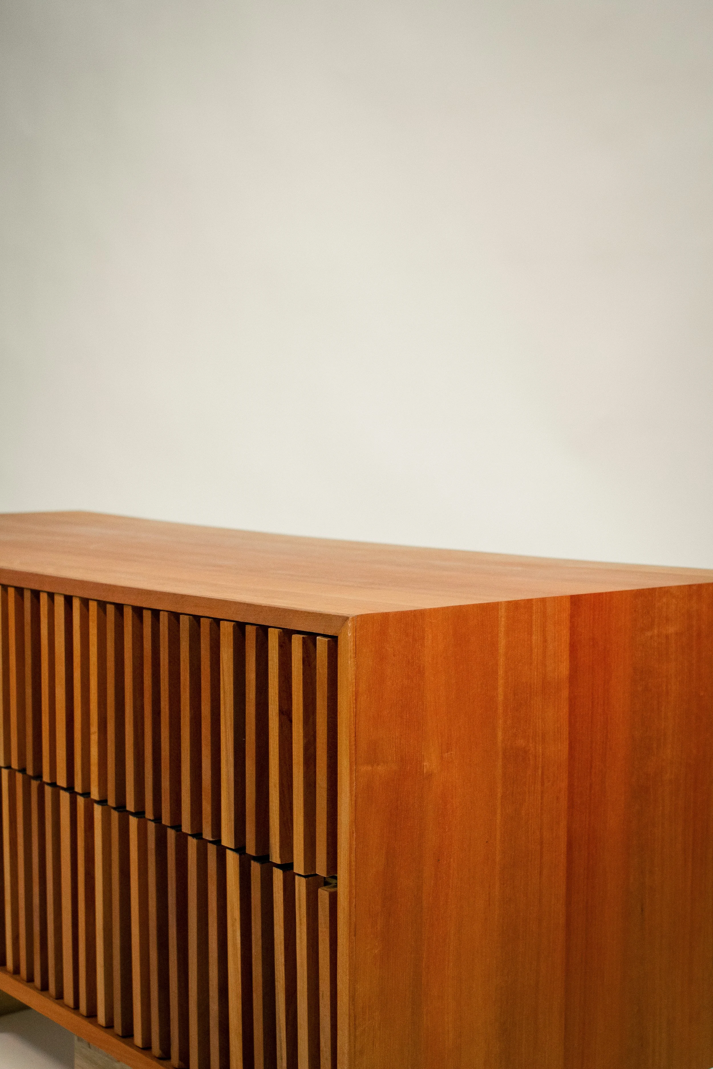 Corner of a wooden piece of furniture with vertical slats, against a plain light-colored wall.