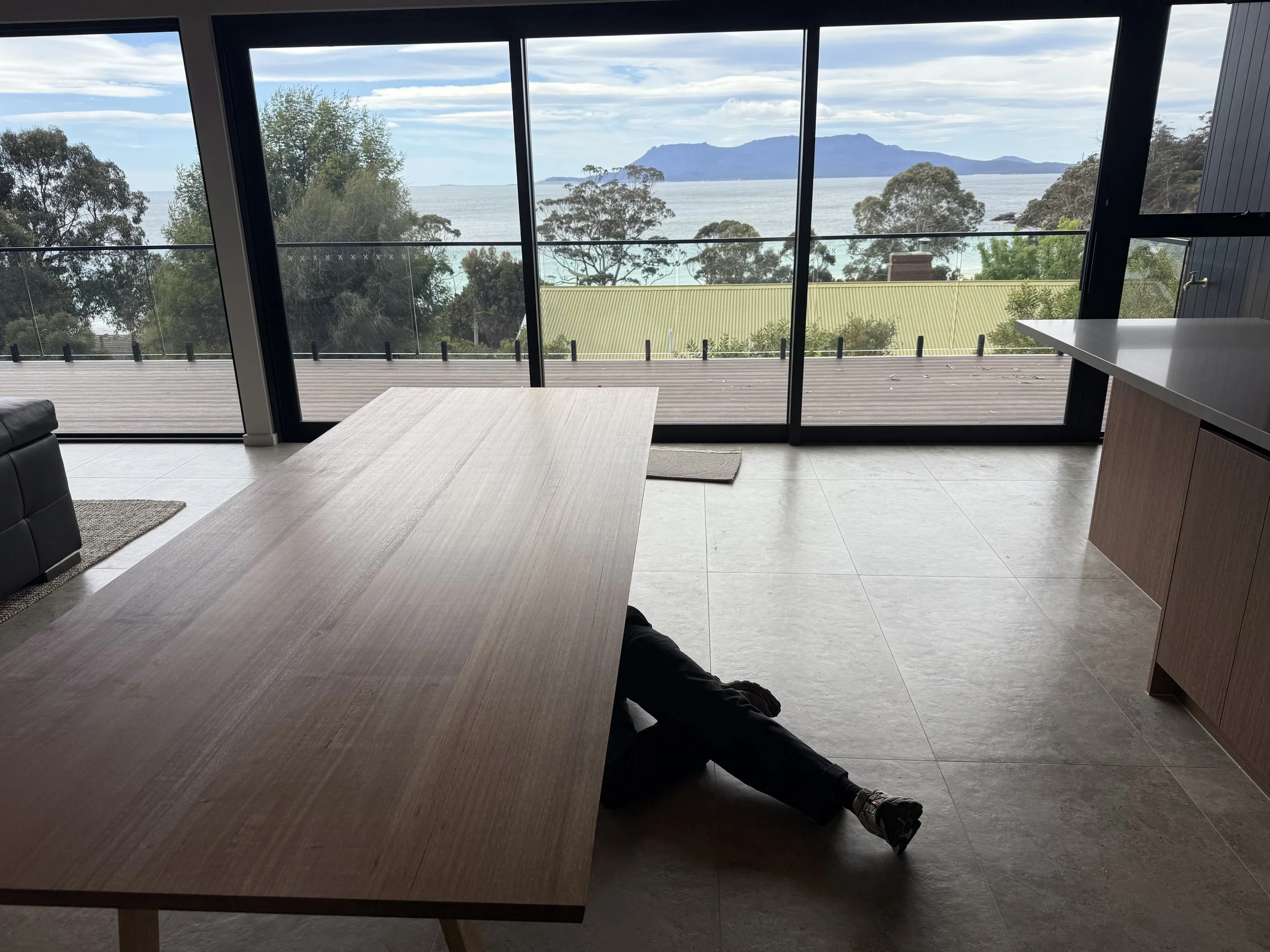 Interior view of a dining room, with a custom dining room table. looking out through large glass sliding doors onto a wooden deck with a scenic view of maria island, ocean and trees under a partly cloudy sky.