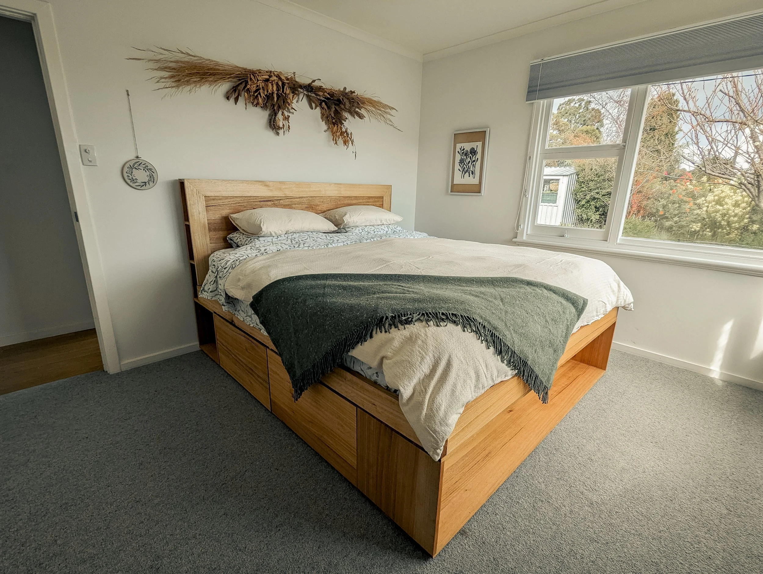 A bedroom with a wooden bed frame, beige bedding, and a dark green throw blanket. There is some wall art above the bed, including a decorative piece made of dried plants and a framed botanical print. The room has large windows revealing trees outside