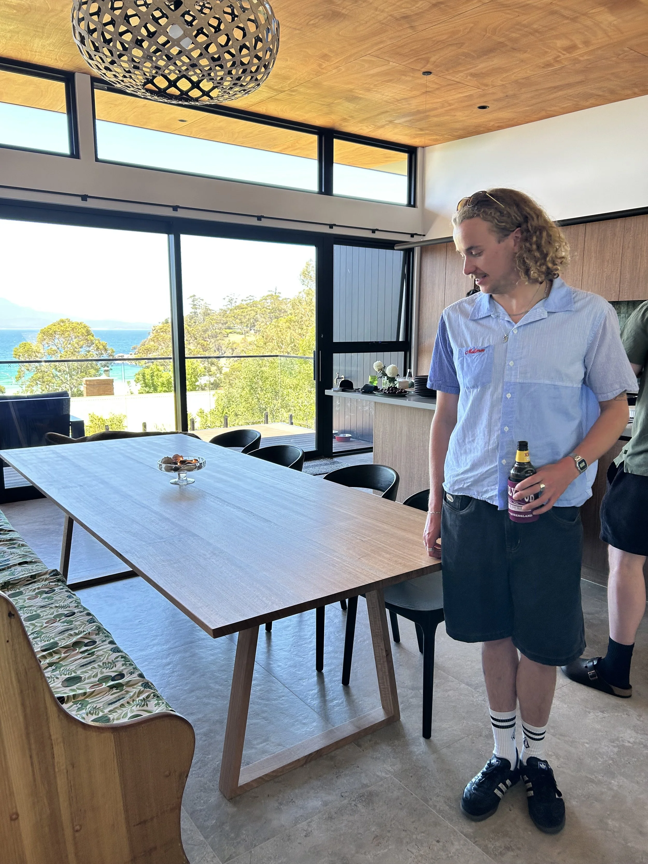 Young man with curly blonde hair wearing a light blue shirt, black shorts, white striped socks, and black sneakers, holding a beer bottle at a dining table in a modern room with large windows showing trees and a body of water outside.