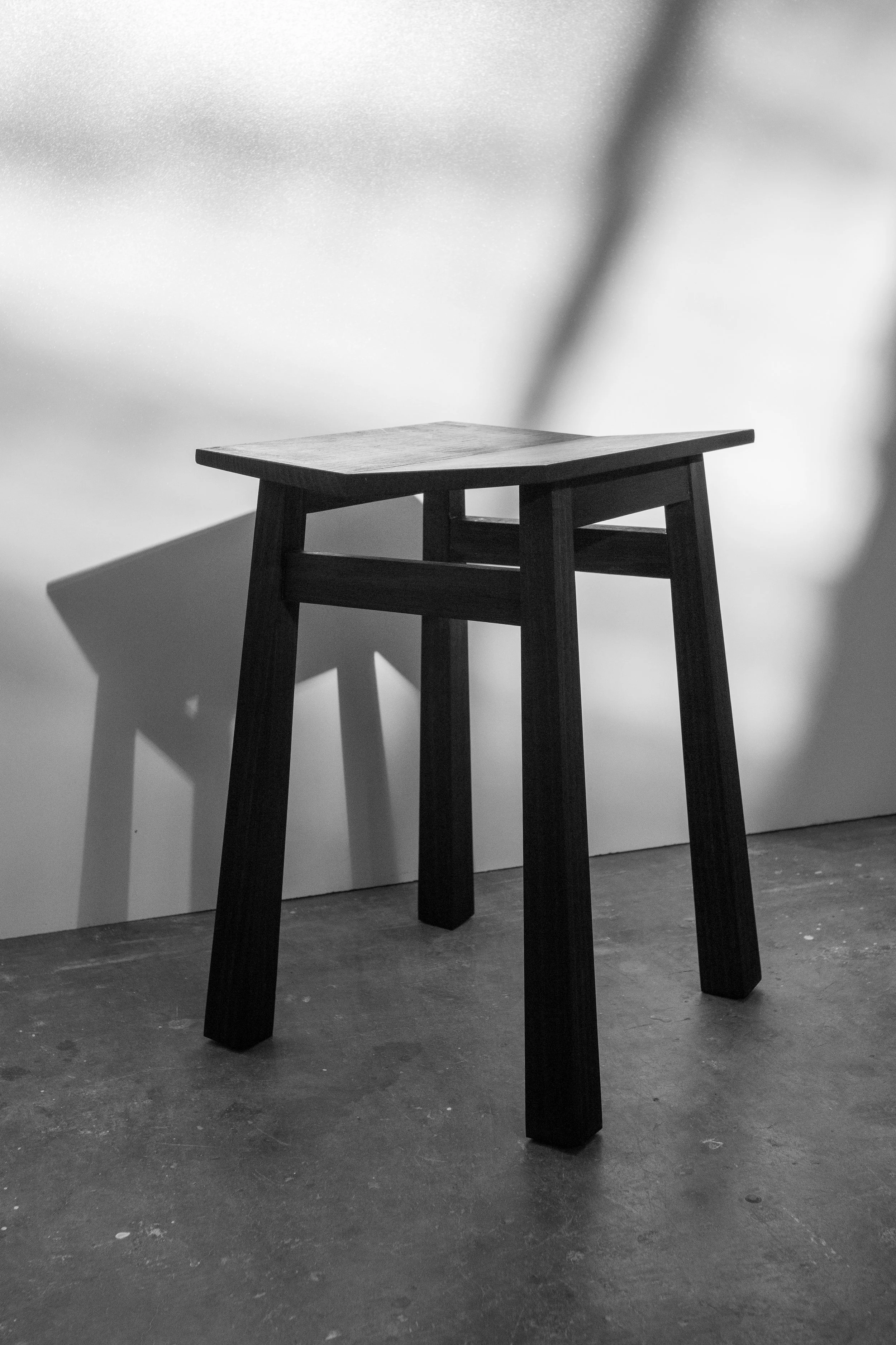 Black and white photo of a wooden stool with a slip in the middle, standing on a concrete floor, against a plain wall.