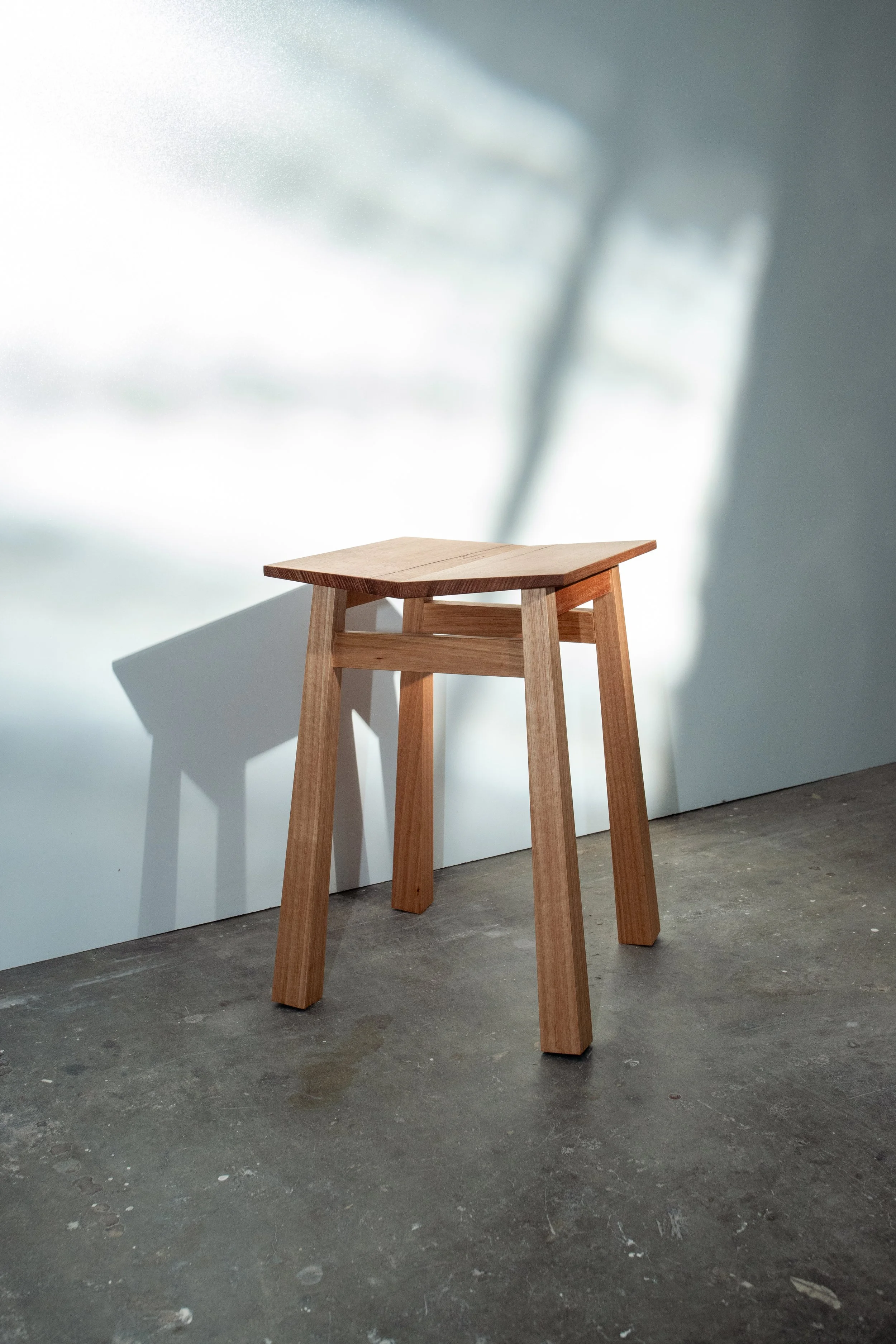 A wooden stool with four legs and a flat top, placed on a concrete floor with public art wall in the background.