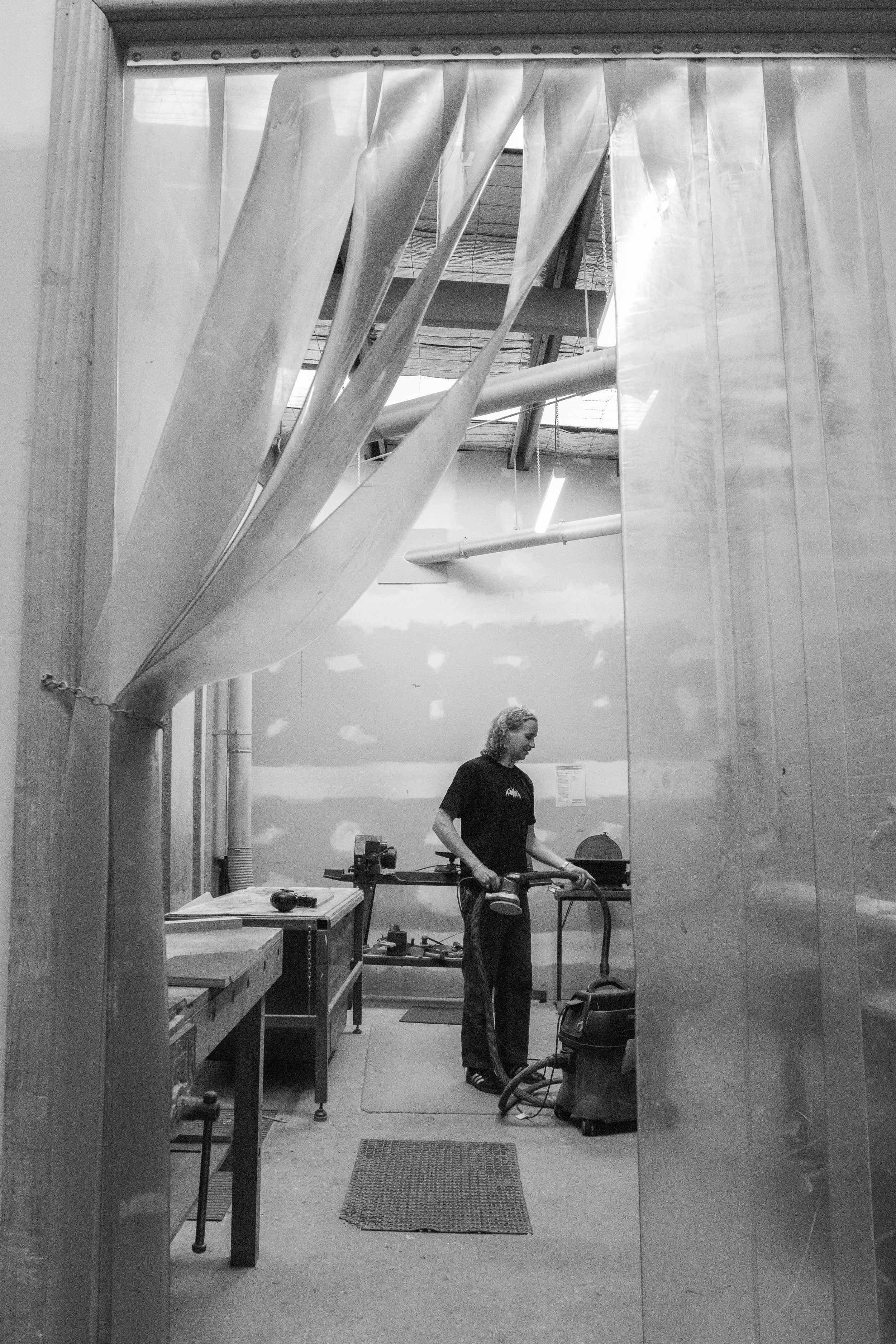 A person standing in a workshop holding an electric sander, with woodworking tools and equipment on tables and shelves around them, viewed through an open doorway.