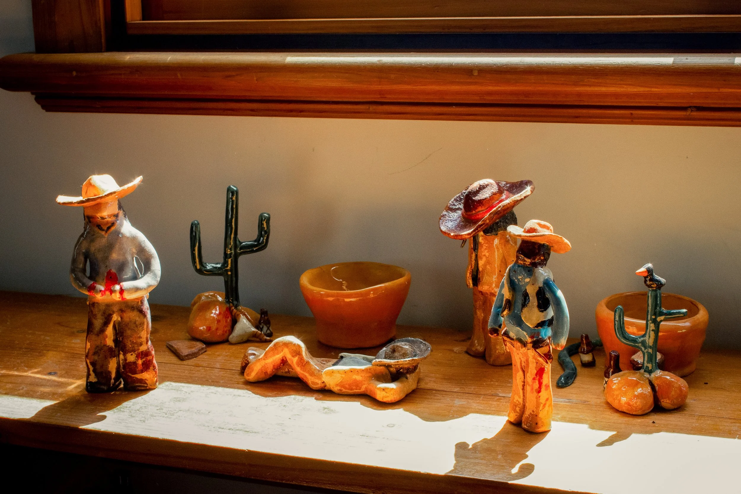 Display of ceramic Mexican-themed figurines including figures wearing sombreros, cacti, and small pots, arranged on a wooden surface with a wooden shelf above.