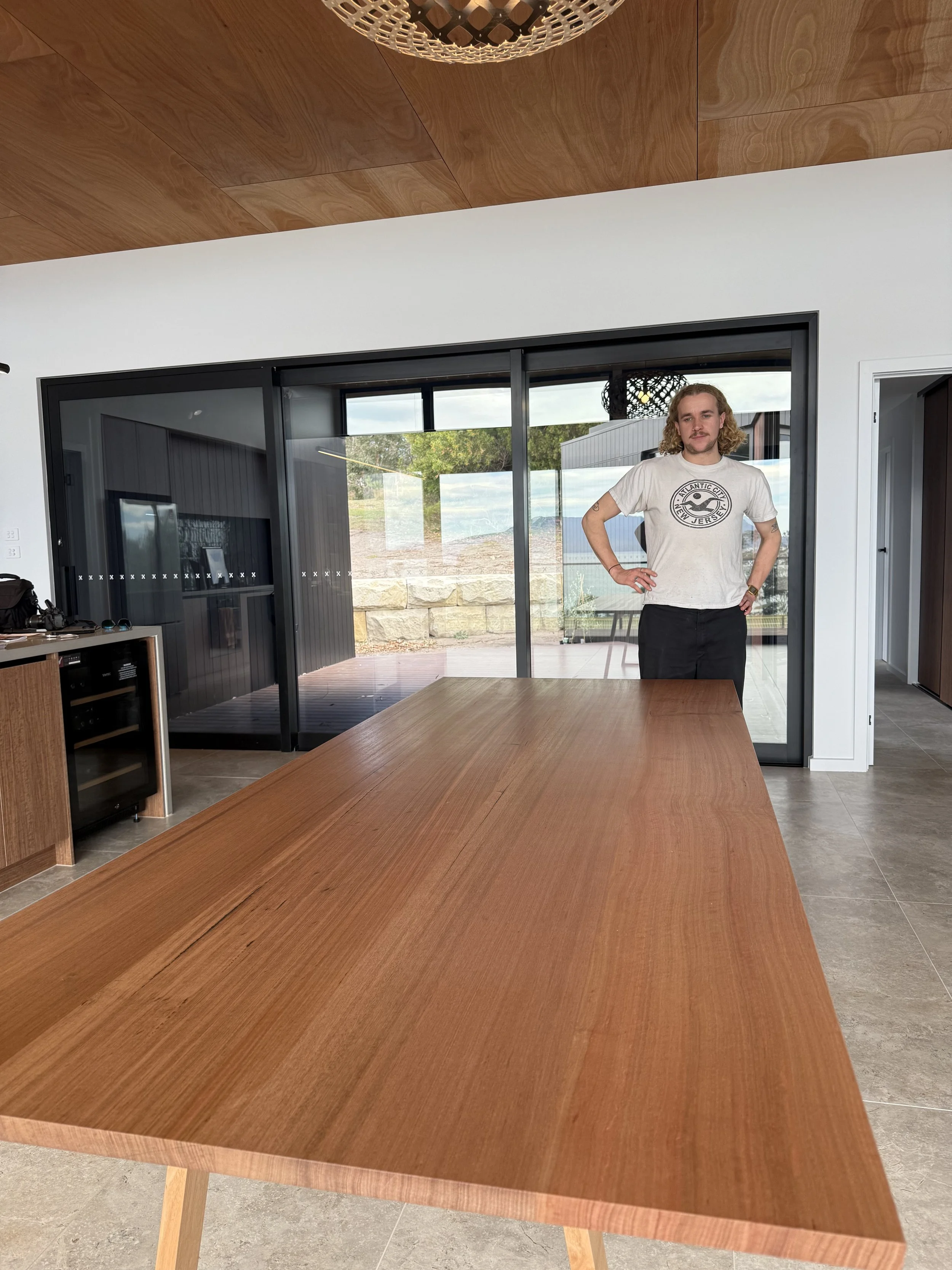 A man with long curly hair wearing a white t-shirt and black pants standing indoors near a large glass door with trees and outdoor scenery visible.