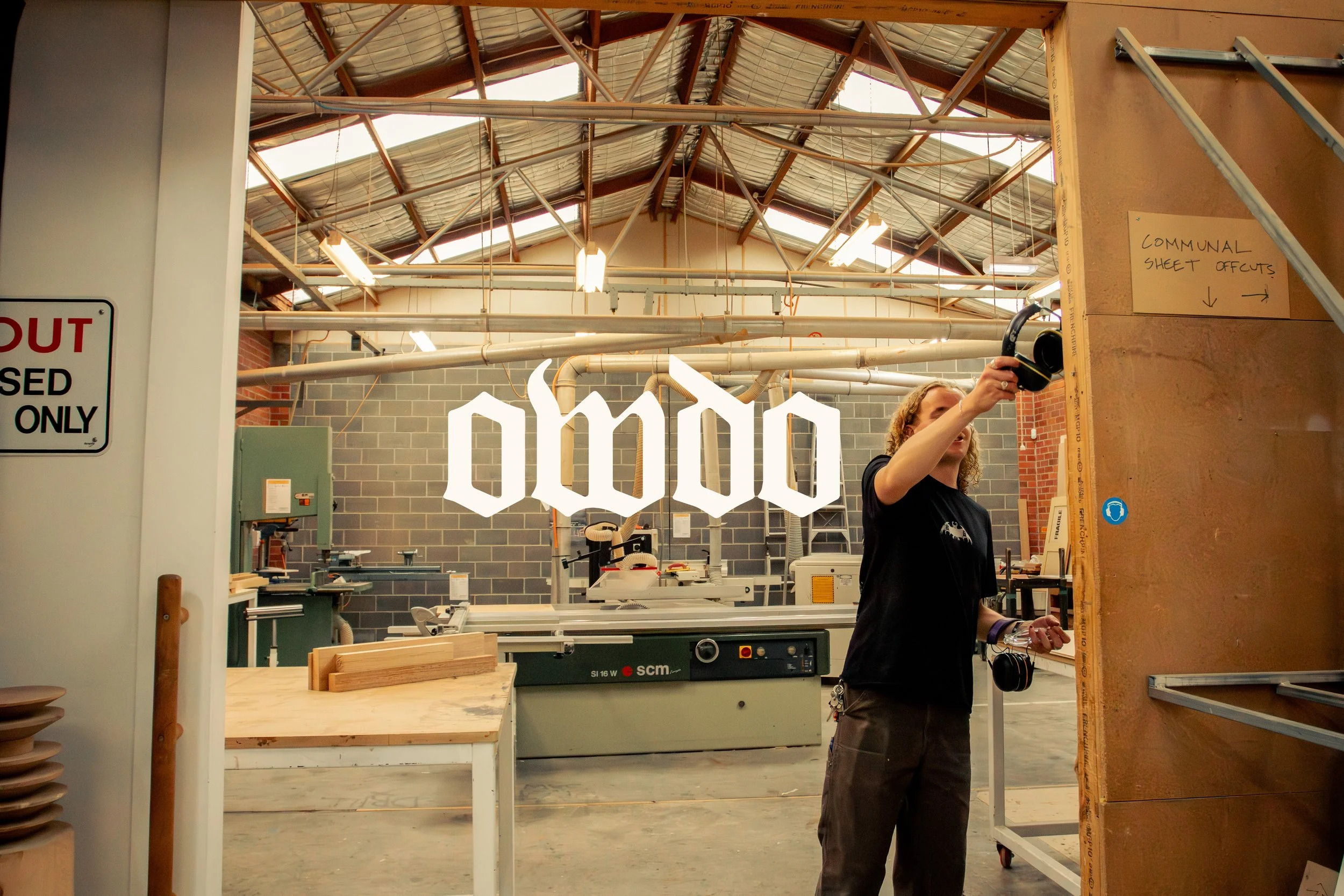 Interior of a workshop with a young man starting work. The workshop has a high ceiling with skylights and various woodworking tools and equipment visible.