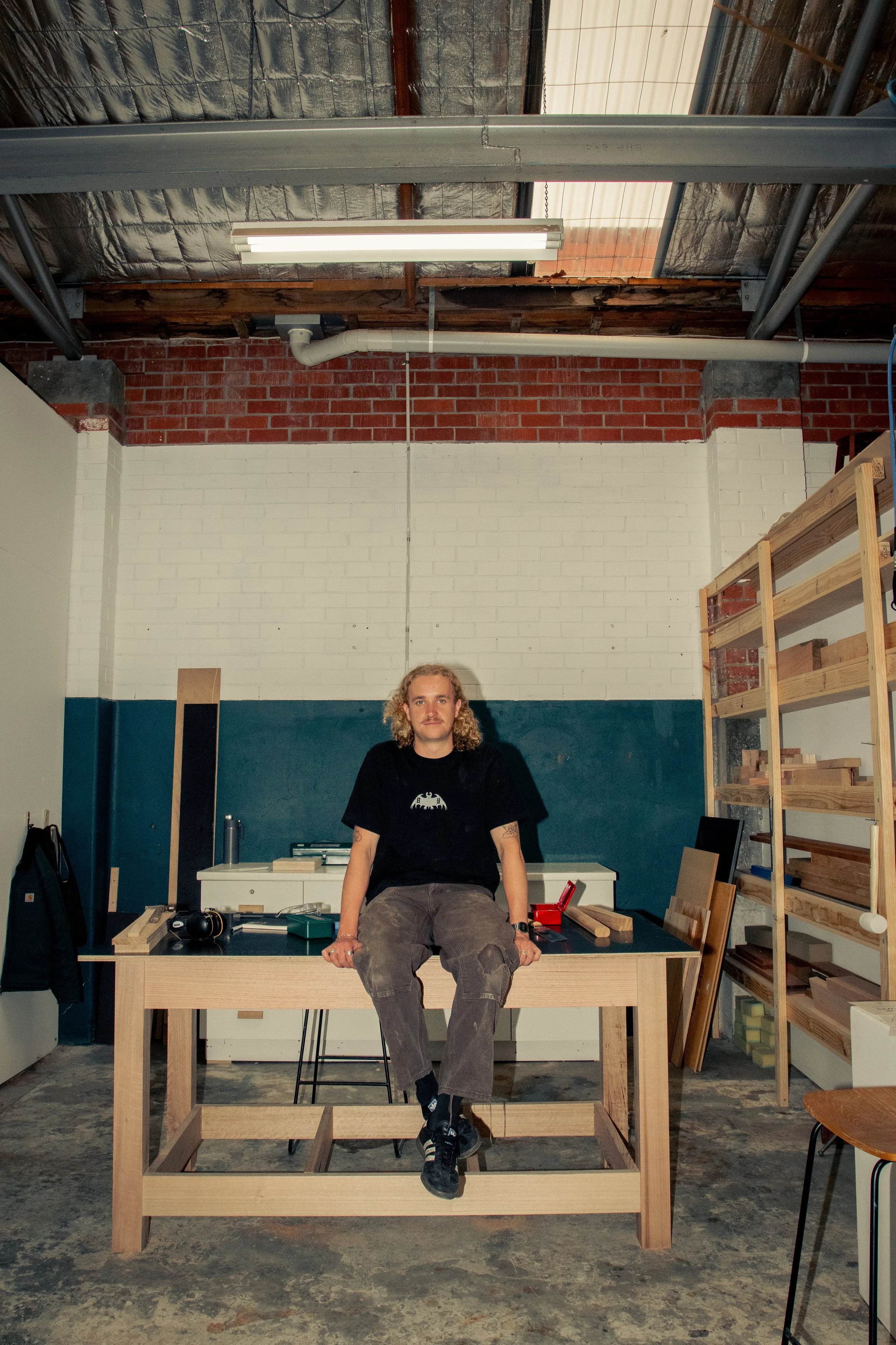 A young man with curly hair and tattoos, sitting on a wooden table in an industrial workspace, surrounded by construction tools, shelves, and unfinished walls.