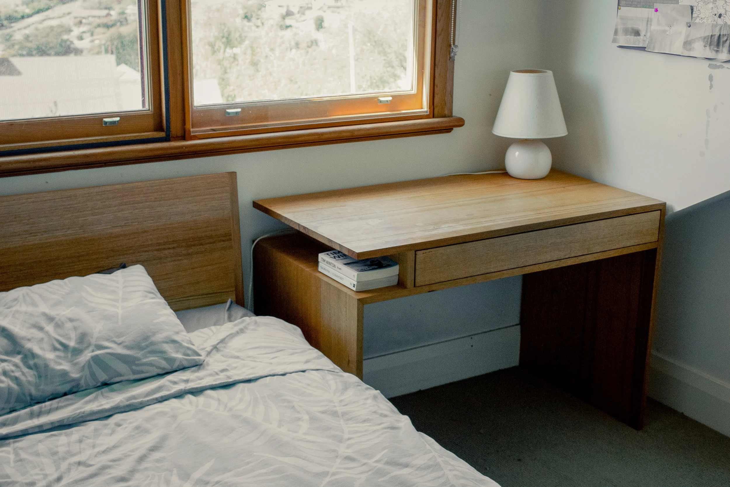 A bedroom with a wooden nightstand and a white table lamp next to a bed with white and blue patterned bedding. There is a window with wooden trim showing trees outside. The wall has some patches of peeling paint and overlapping papers.
