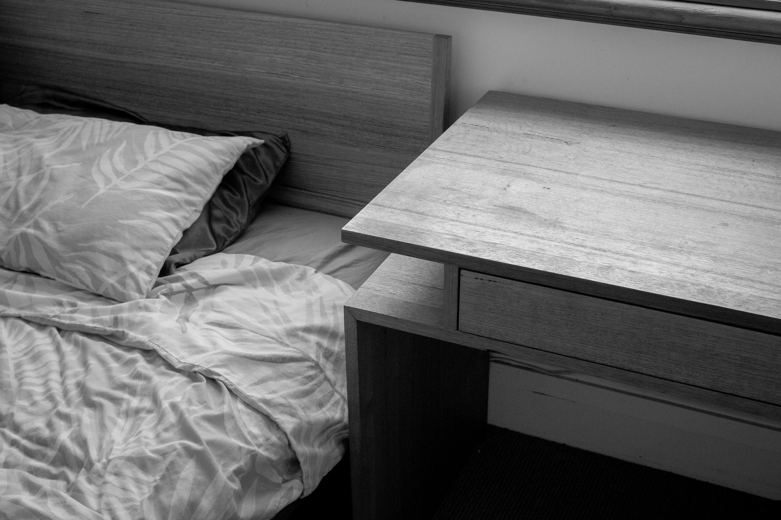 A bed with crumpled floral patterned sheets and pillows next to a wooden nightstand, with a wooden headboard in a monochrome color scheme.