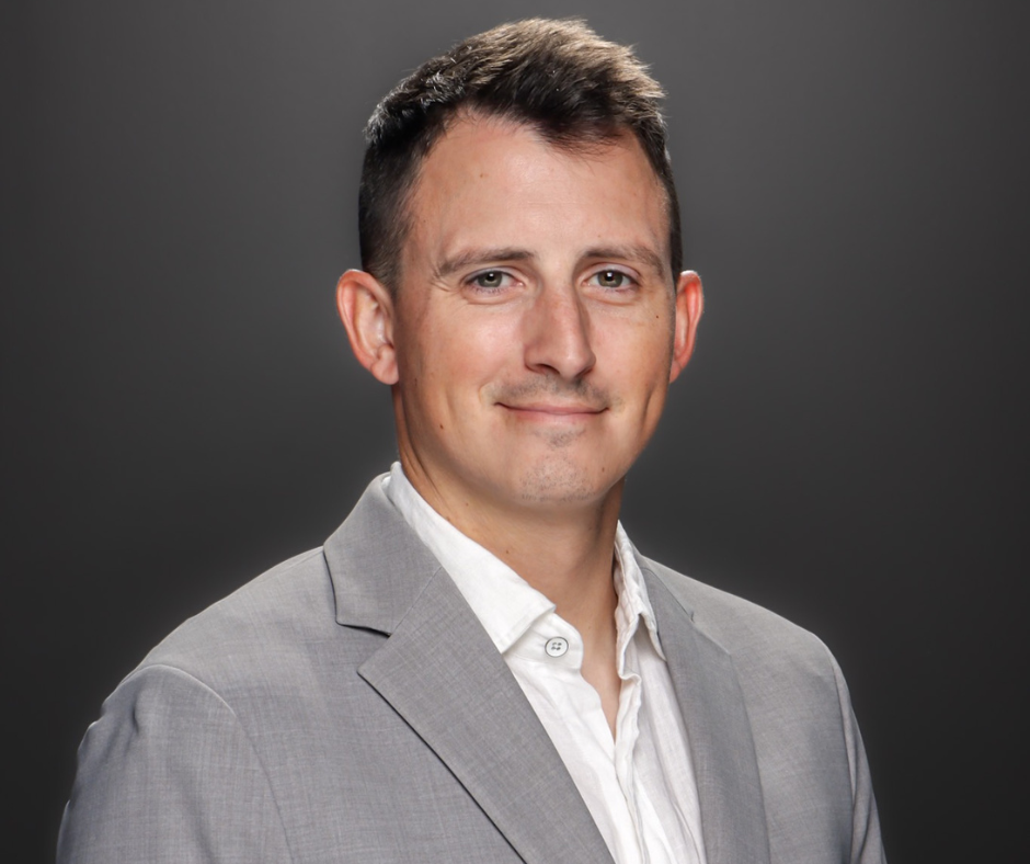 Professional headshot of a man with short dark hair, wearing a light gray blazer and a white shirt, smiling slightly against a dark gray background.