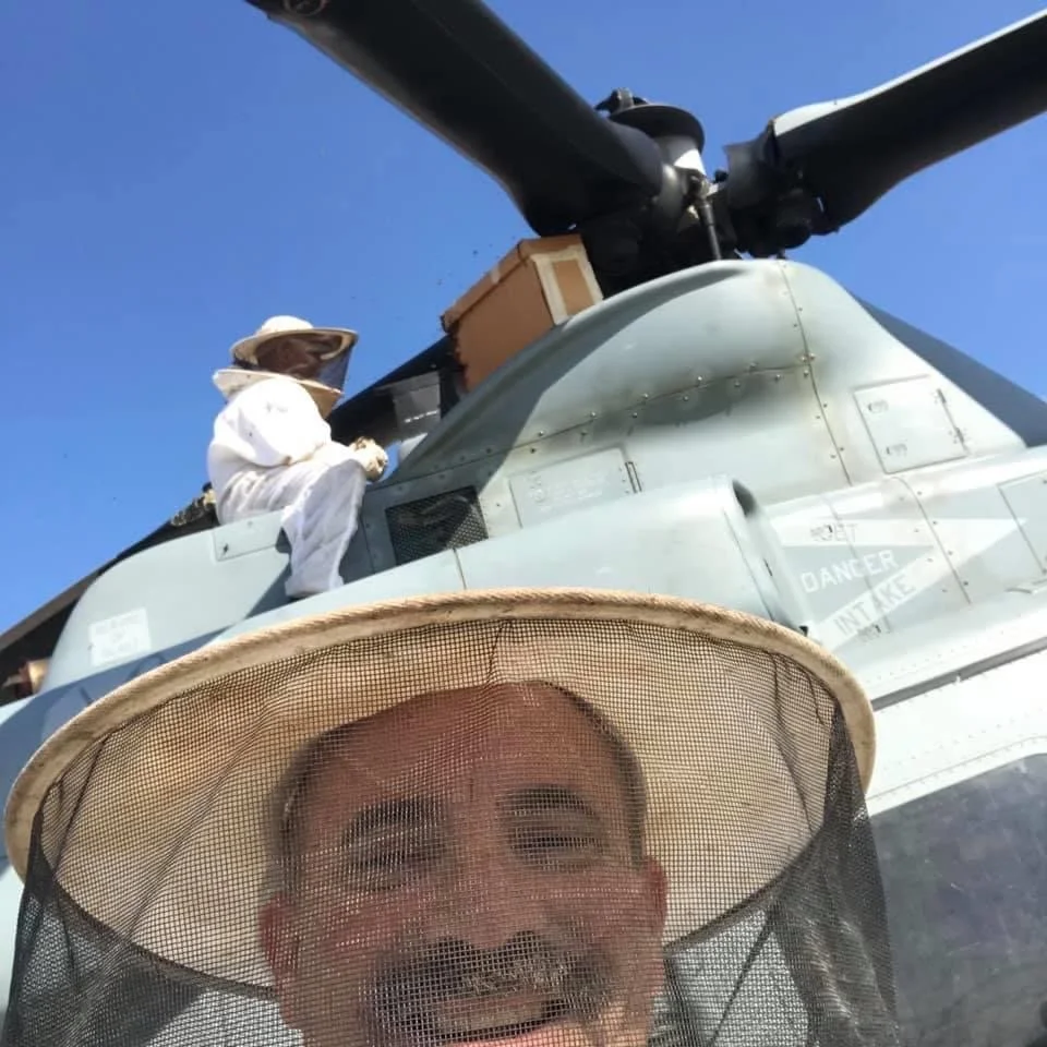 Man with a beard smiling inside a beekeeping veil in front of a military helicopter with a person inside the cockpit.