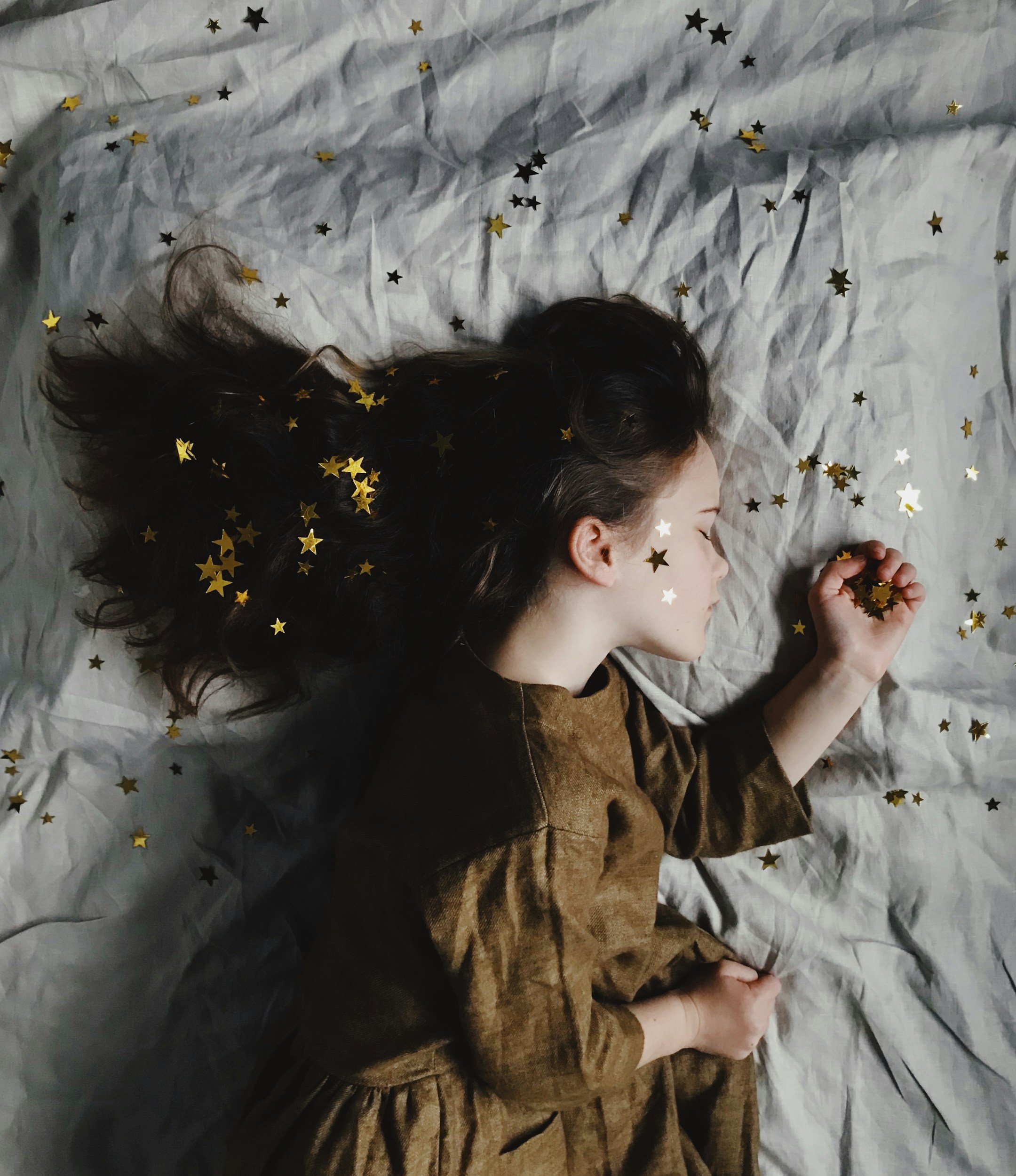 A woman with dark, curly hair lying on a bed covered with white sheets decorated with black and gold star-shaped confetti, with her eyes closed and hands clasped.