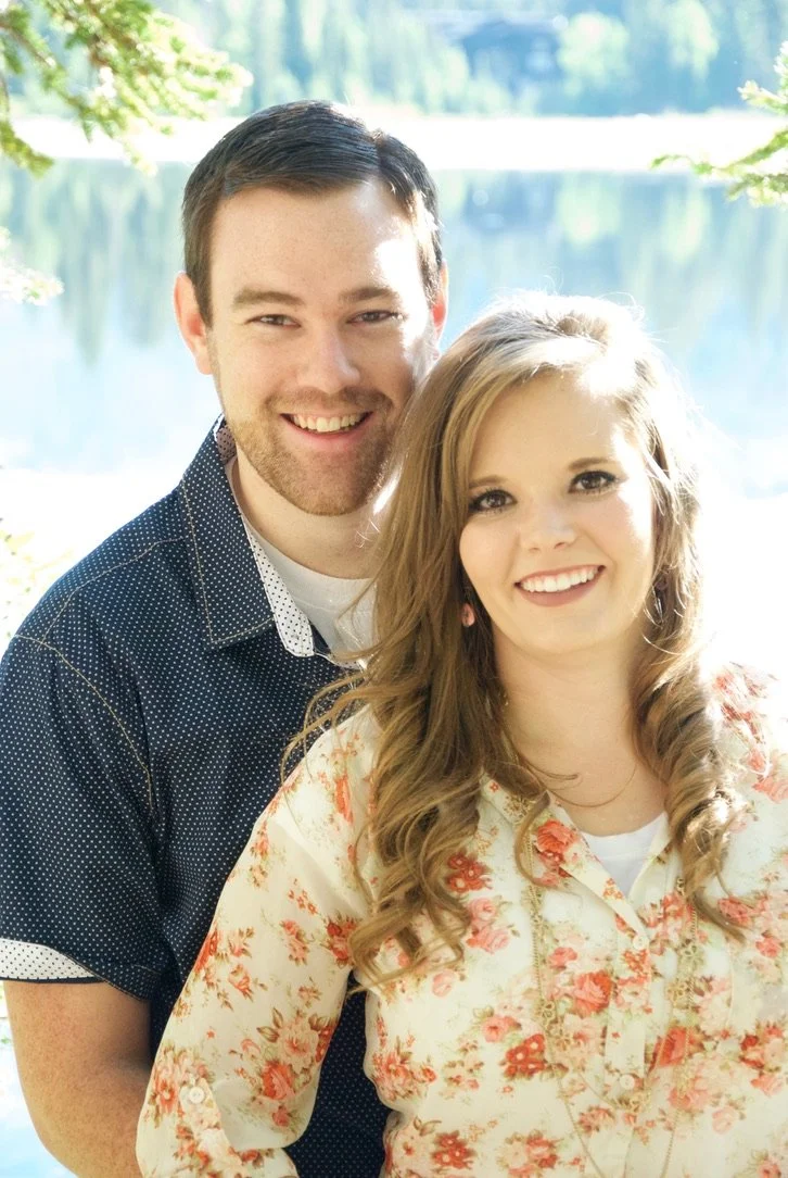 A smiling couple standing outdoors near a lake with trees in the background. The man has short dark hair and a beard, wearing a dark button-up shirt. The woman has long wavy blonde hair, wearing a light-colored floral blouse.