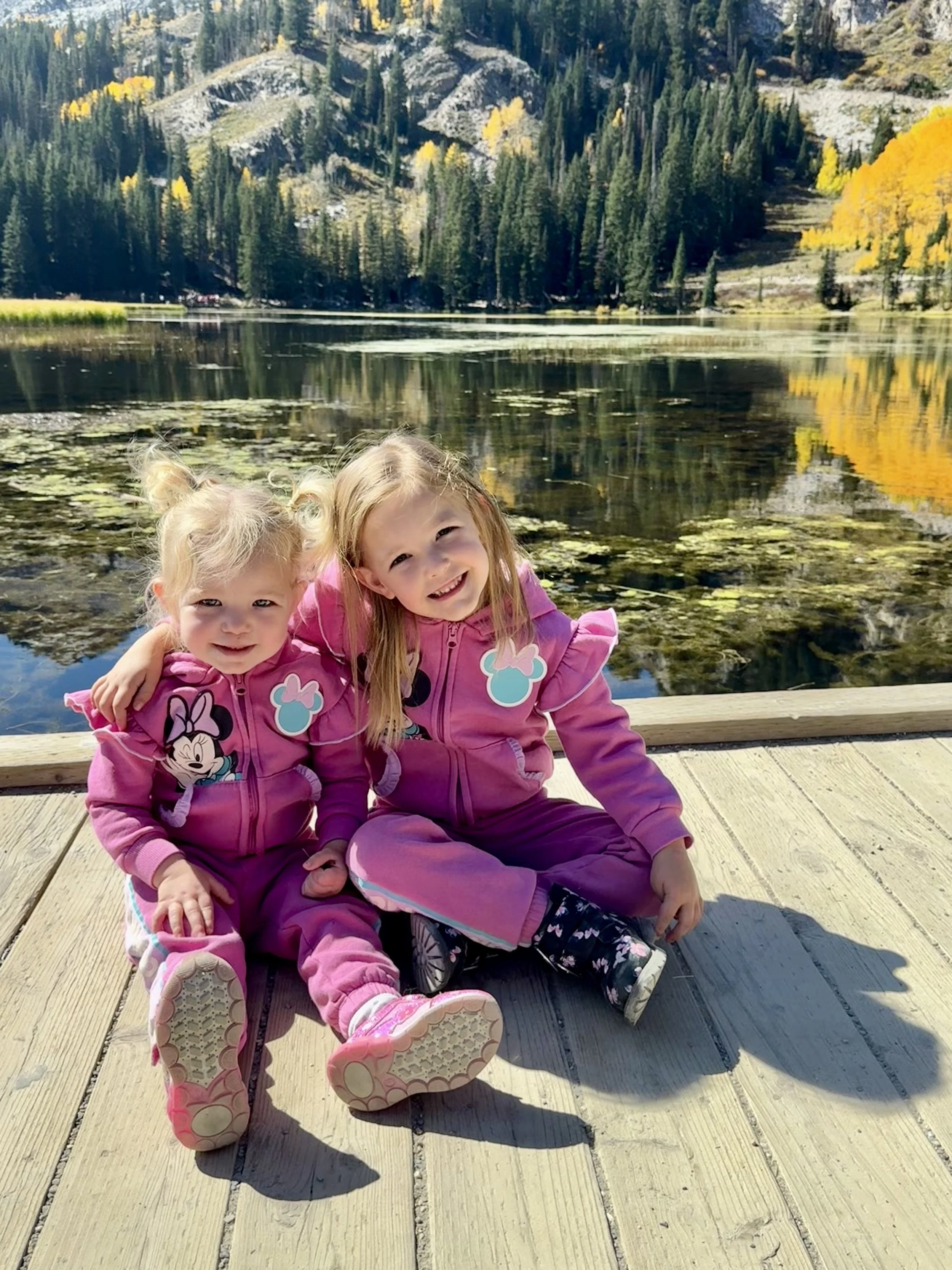 Two young girls sitting on a wooden dock by a lake with mountains and evergreen trees in the background, wearing matching pink outfits with Minnie Mouse and bunny designs, enjoying a sunny day.