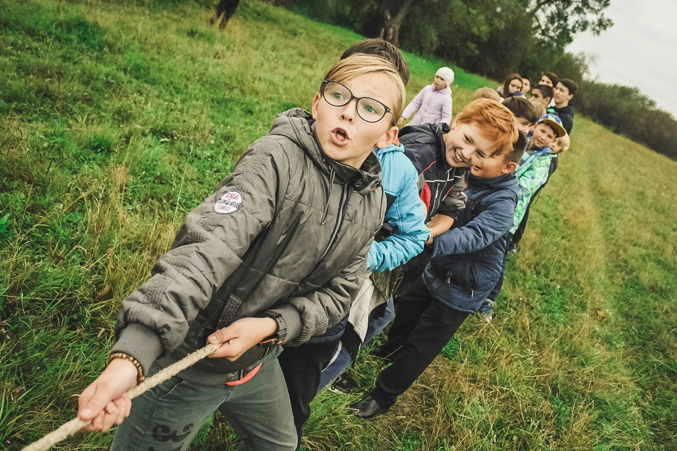 A group of children holding a rope, standing in a line outdoors on a grassy hill, with trees in the background, participating in an outdoor activity.