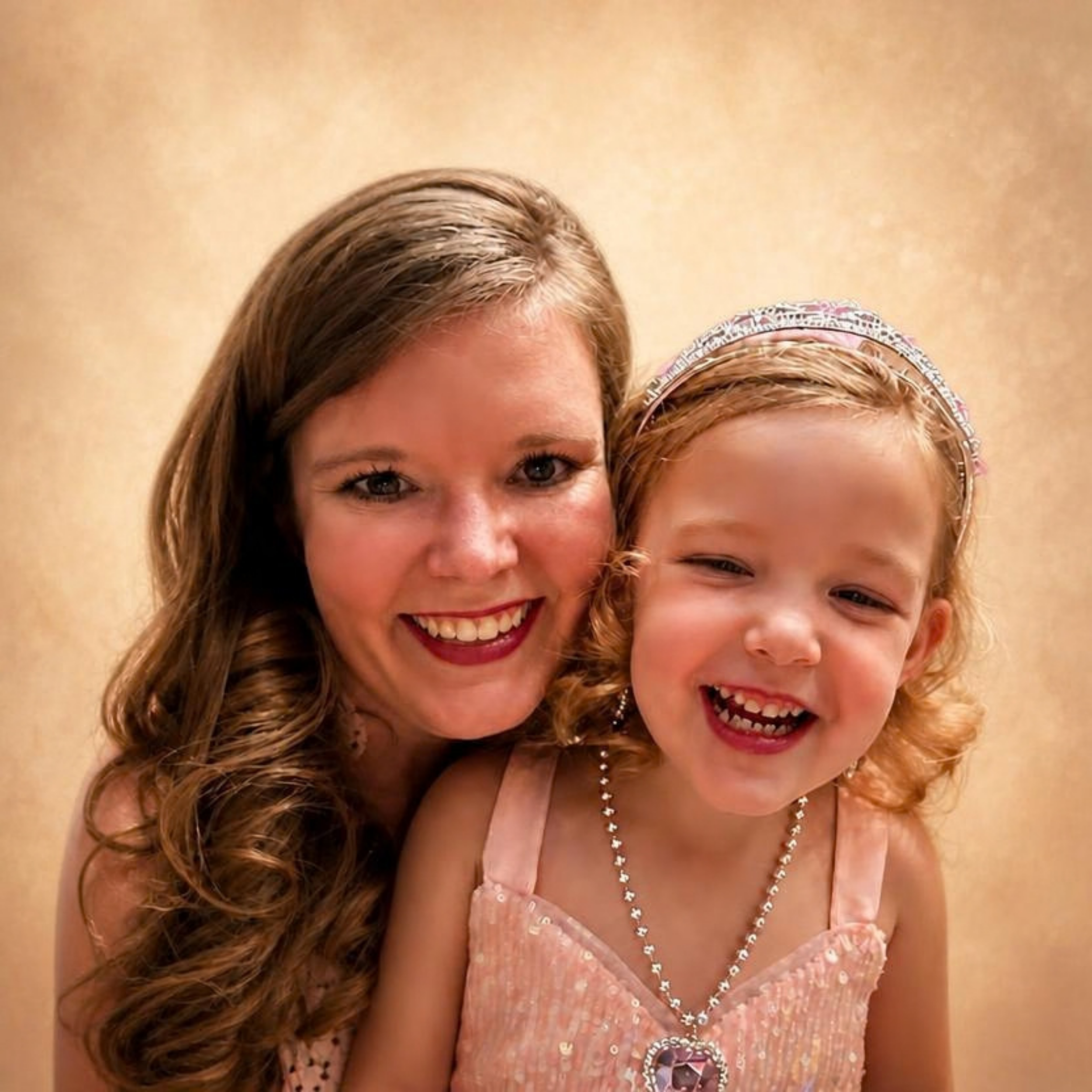 Close-up of two smiling women, one with long wavy hair and the other with curly red hair, dressed in elegant pink and pearl attire, wearing necklaces, earrings, and a decorative headband, against a plain background.