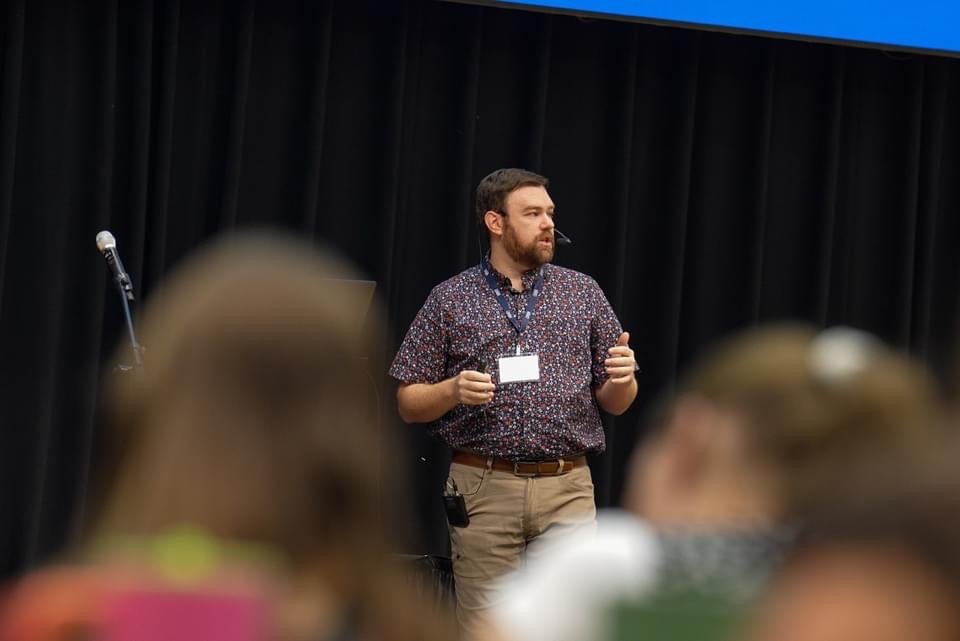 A man with a beard and short brown hair, wearing a patterned short-sleeve shirt, khaki pants, and a lanyard, standing on a stage with a microphone on a stand behind him. The background is black curtains, and blurred audience members are in the foreground.