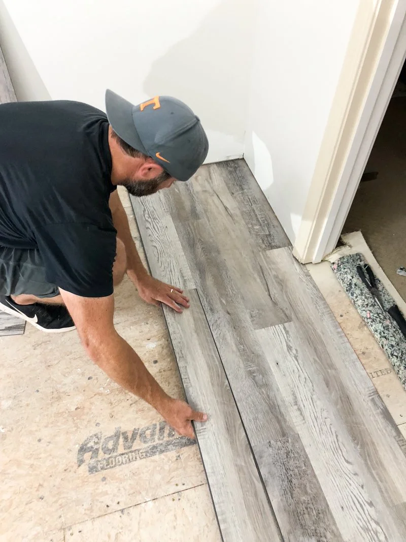 A person installing LVT or wood-look tile flooring in a room, kneeling on the floor, wearing a gray cap, black shirt, and gray shorts.