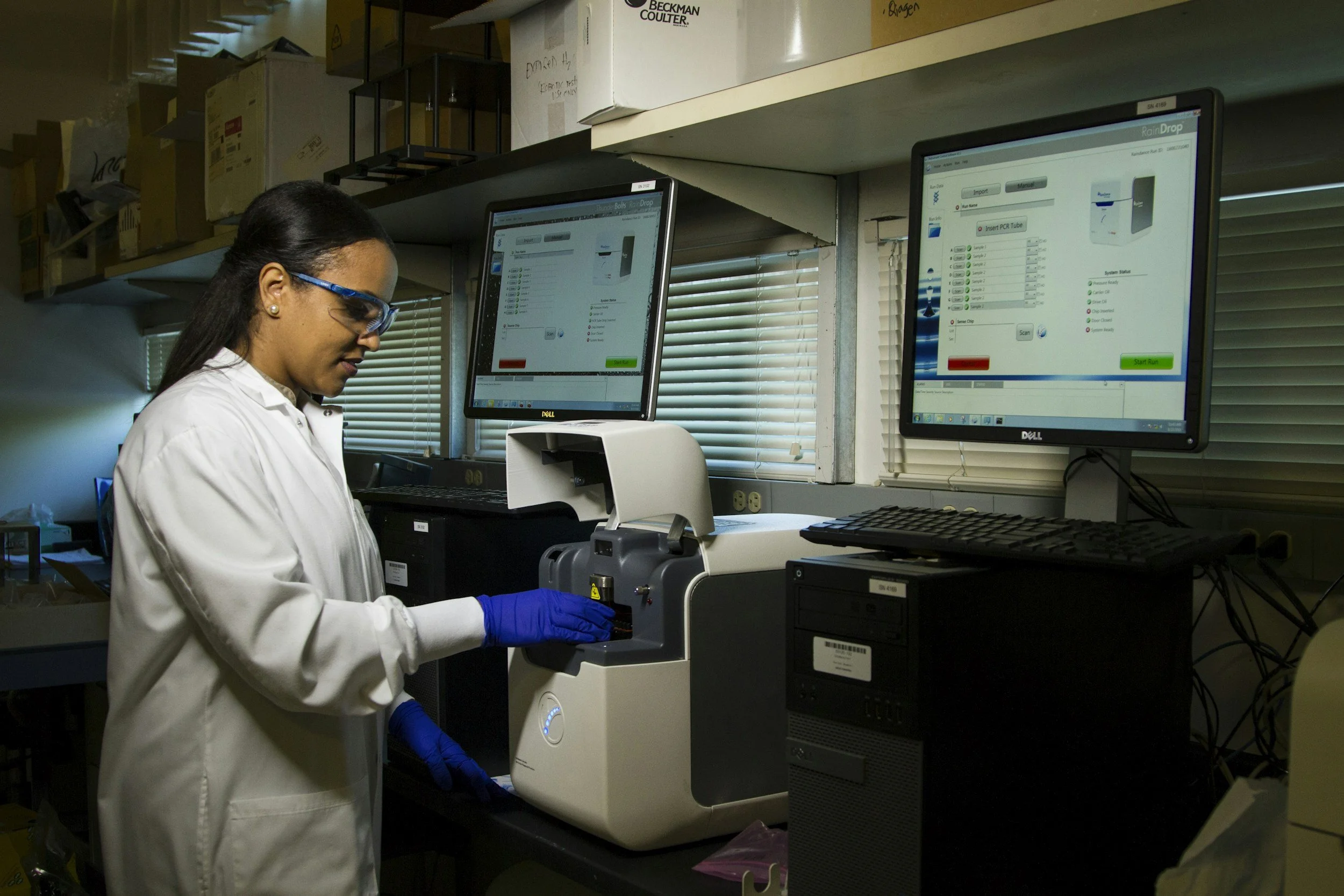 A scientist in a white lab coat and blue gloves working with laboratory equipment in a laboratory setting. There are two computer monitors on the desk displaying lab software, and shelves with boxes and supplies behind her.
