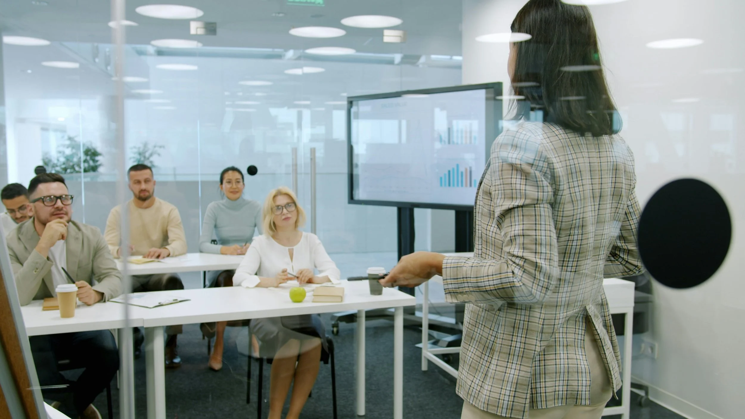 A woman presenting in front of a group of colleagues in a conference room with a large screen displaying graphs.