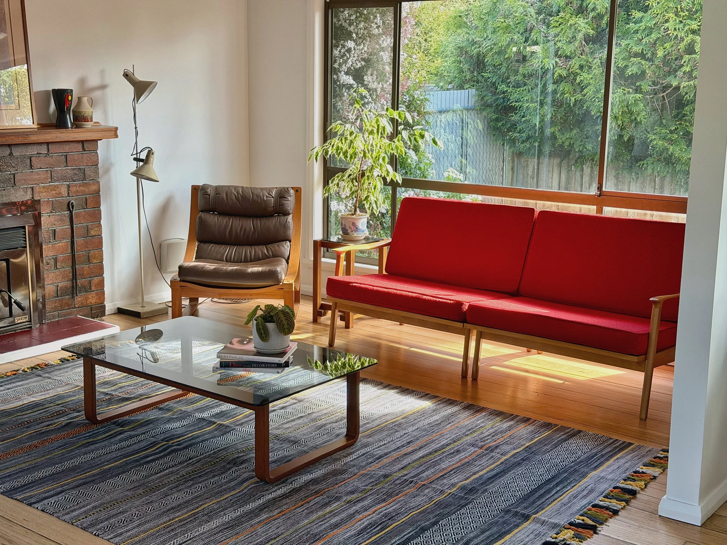 Living room with brick fireplace, brown cushioned armchair, red cushioned sofa, glass coffee table with a plant, and large windows showing greenery outside.