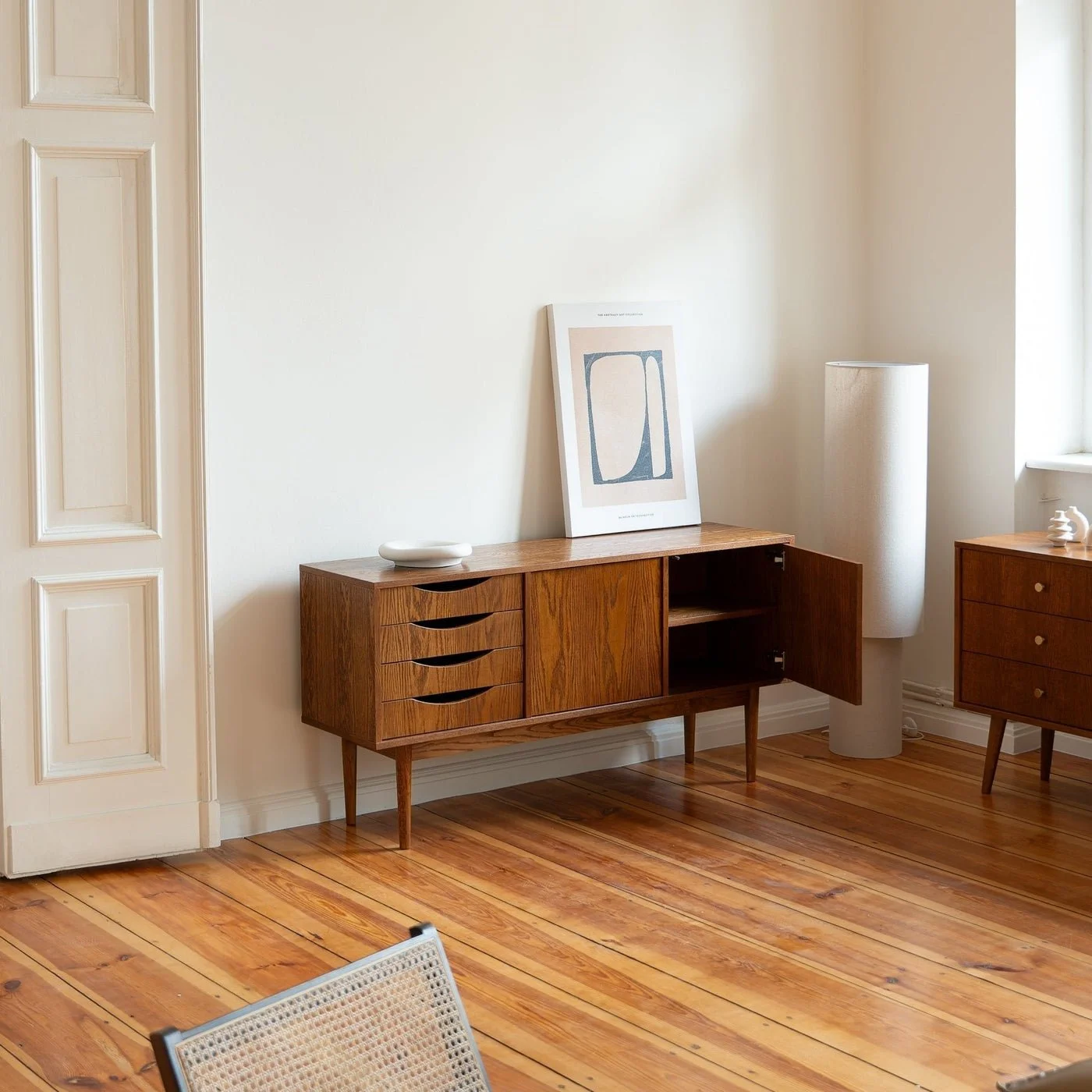 Mid-century modern wooden sideboard with four drawers, a cabinet, a piece of abstract art, a white ceramic dish, and a tall white floor lamp in a bright room with white walls and wooden floors.