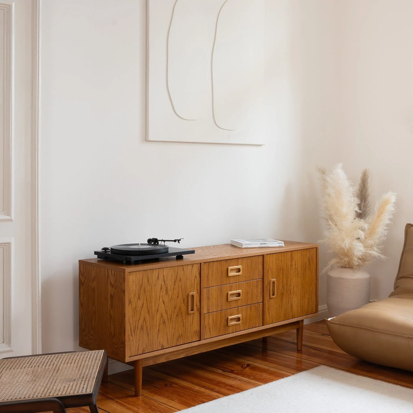 A wooden sideboard with a record player and a book on top, next to a beige chair and a vase with pampas grass in a living room with white walls and hardwood flooring.