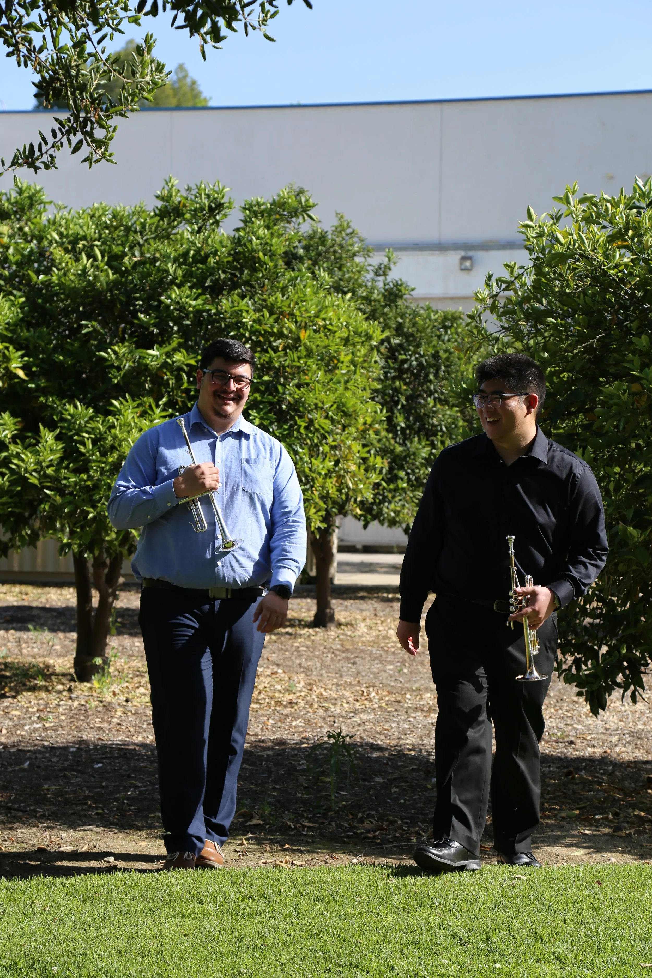 Two men are walking outdoors in a garden with green bushes, carrying trumpets and smiling. One wears a blue shirt and glasses, the other wears a black shirt and glasses.