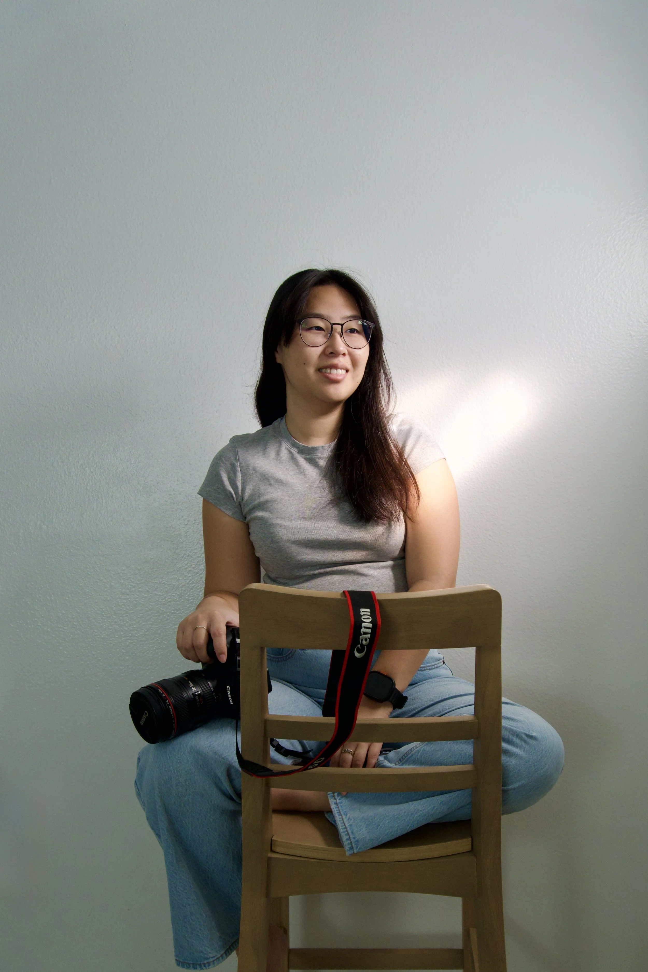 A woman with black hair and glasses sits cross-legged on a wooden chair against a plain white wall. She is holding a Canon camera with a strap resting on the back of the chair, and is wearing a light gray T-shirt and blue jeans.