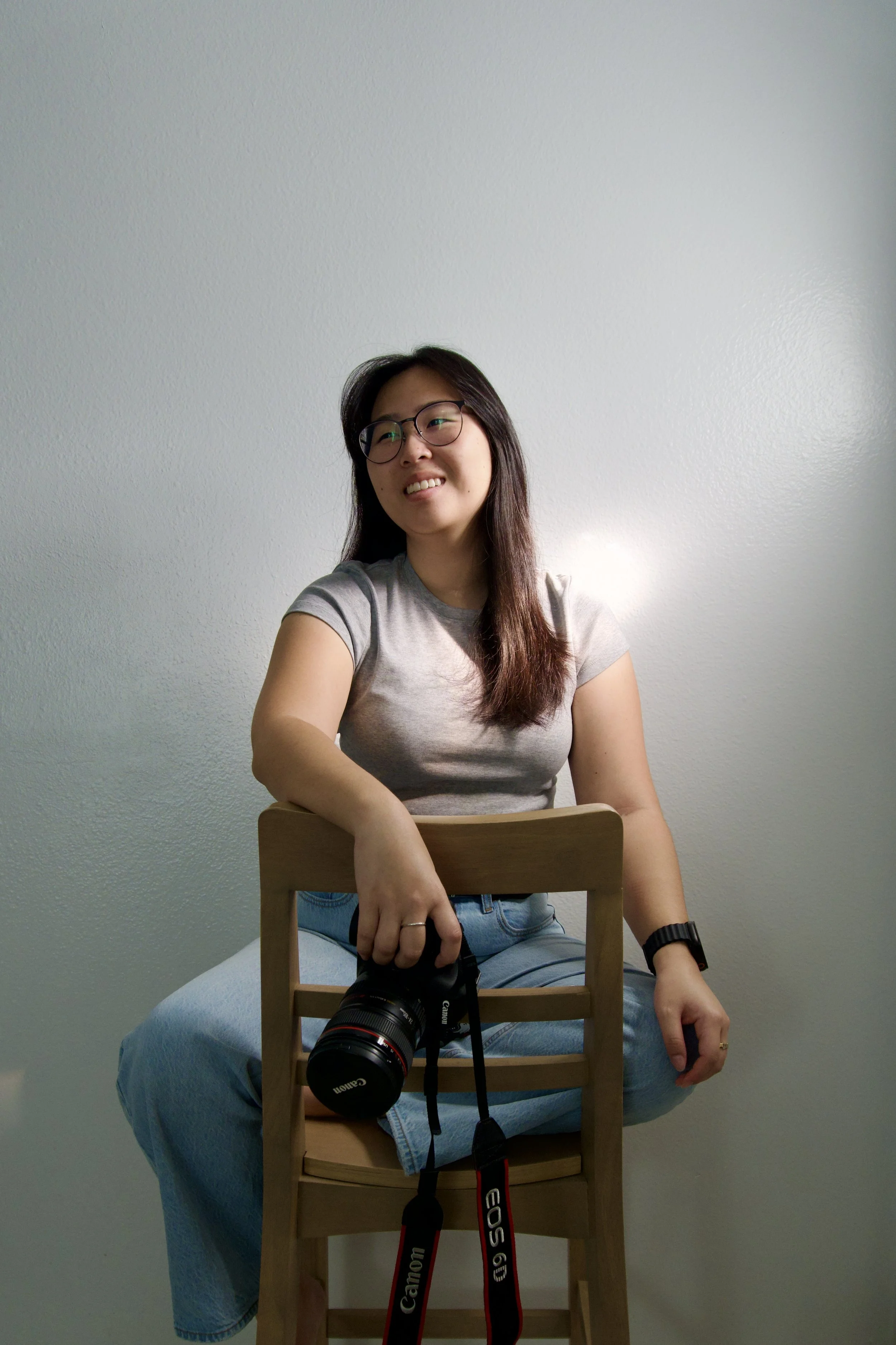 A young woman with long dark hair, glasses, and a grey t-shirt sitting on a wooden chair against a plain light-colored wall, holding a Canon camera.