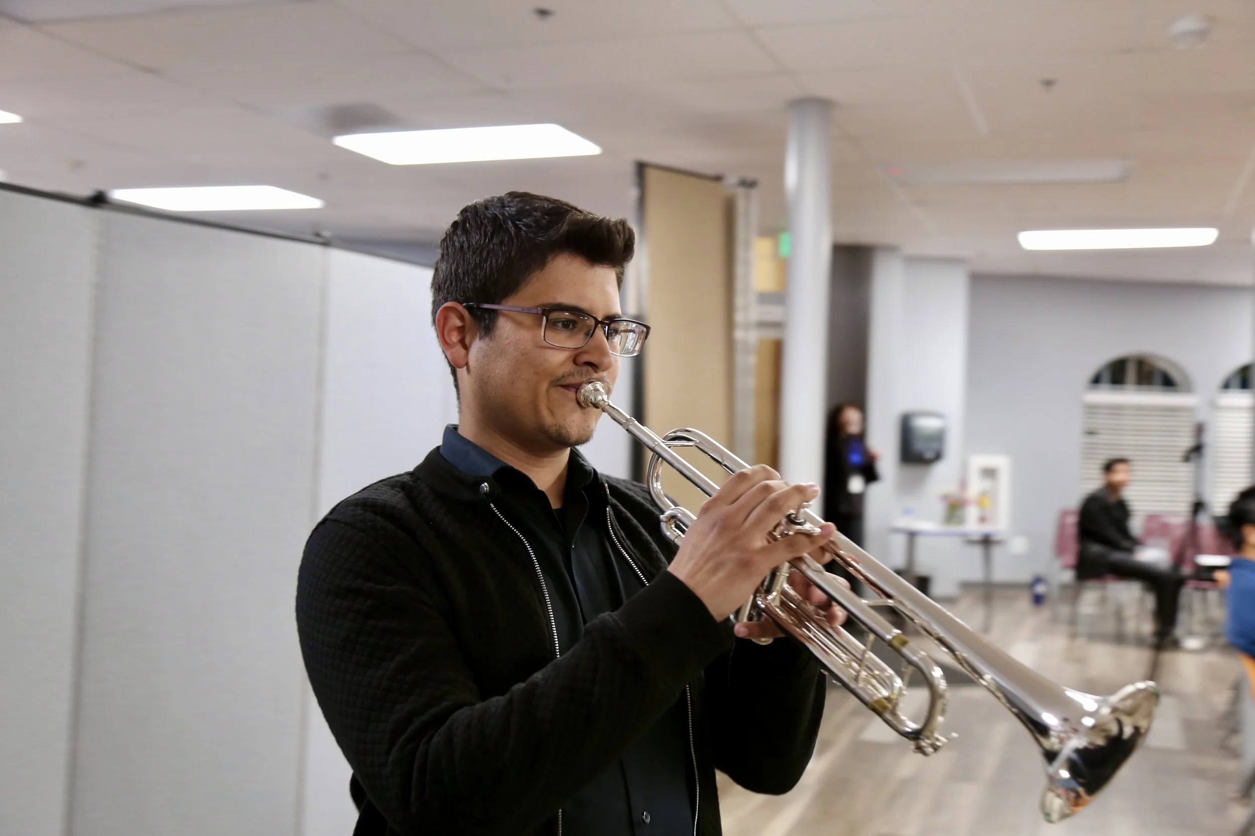 A young man with glasses playing a silver trumpet indoors with a focused expression.