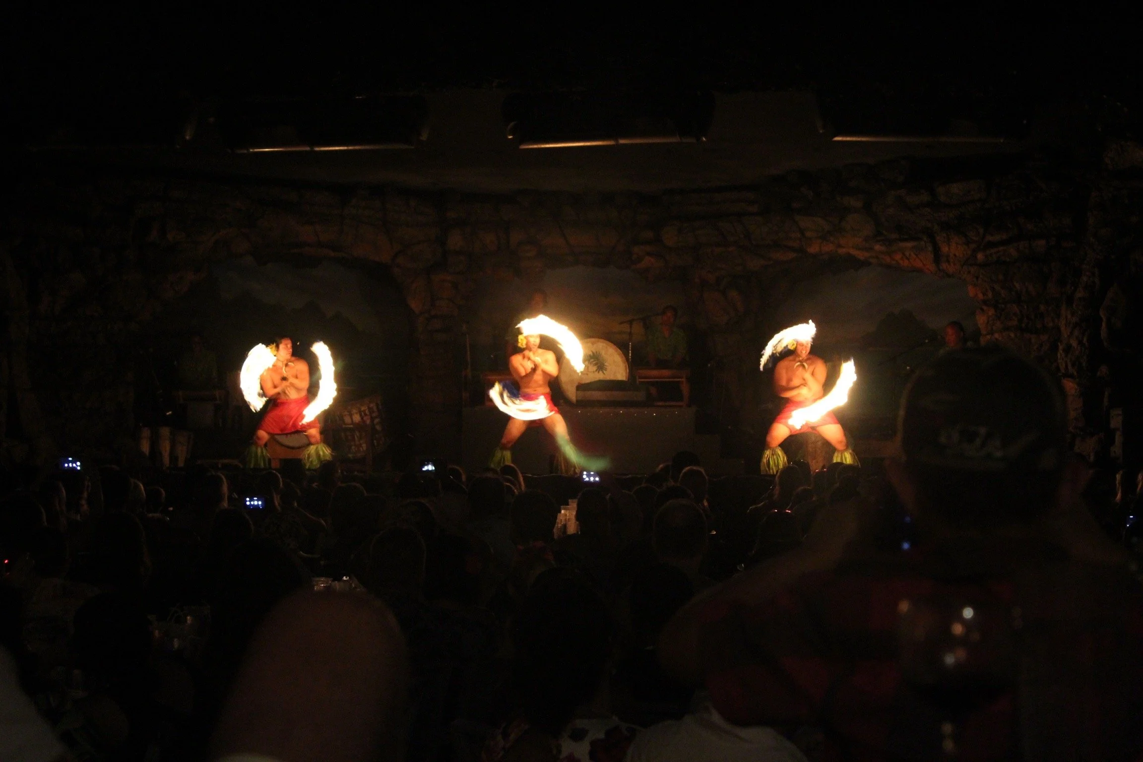Three fire dancers performing on stage at night with stone arch background, audience watching and some holding up phones.